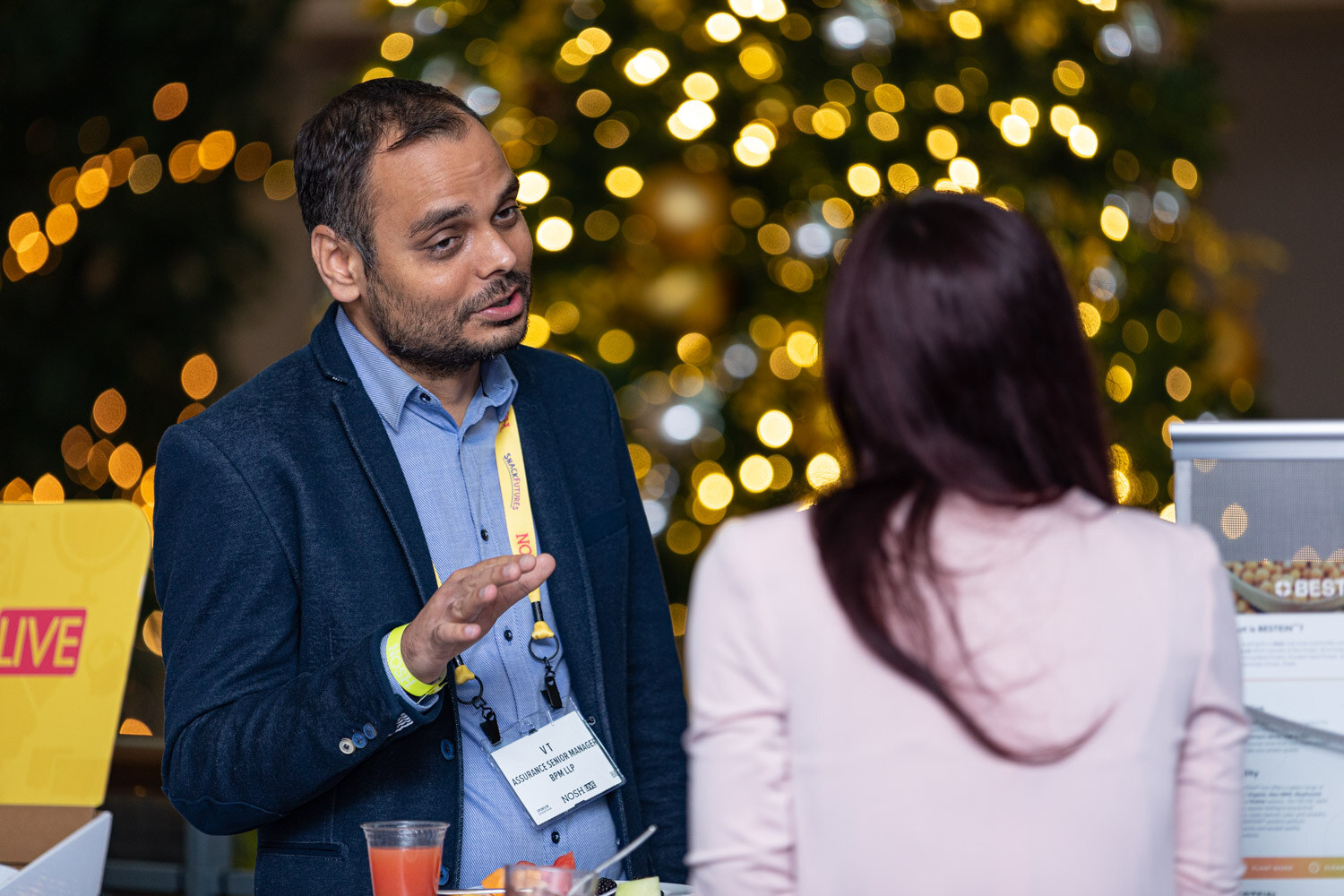 Man in a navy blazer over a light blue shirt with a Nosh lanyard gesturing as he talks to a woman with long dark hair seen from behind, with a lit tree behind them.