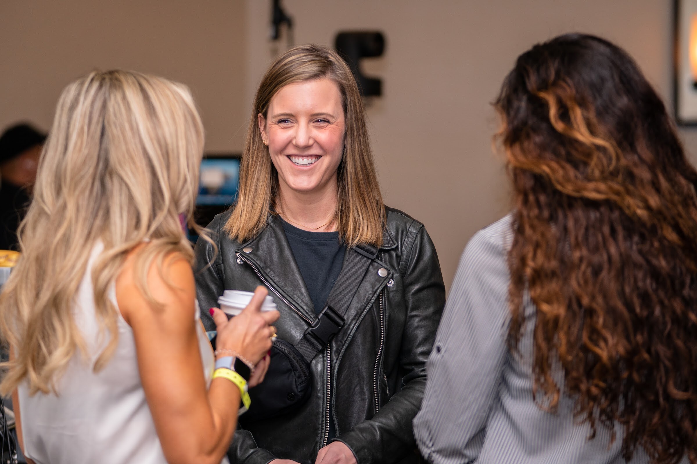 Woman with straight light brown hair in a black leather jacket holding a coffee cup and laughing, facing a woman with long blonde hair and a woman with long curly hair seen from behind.