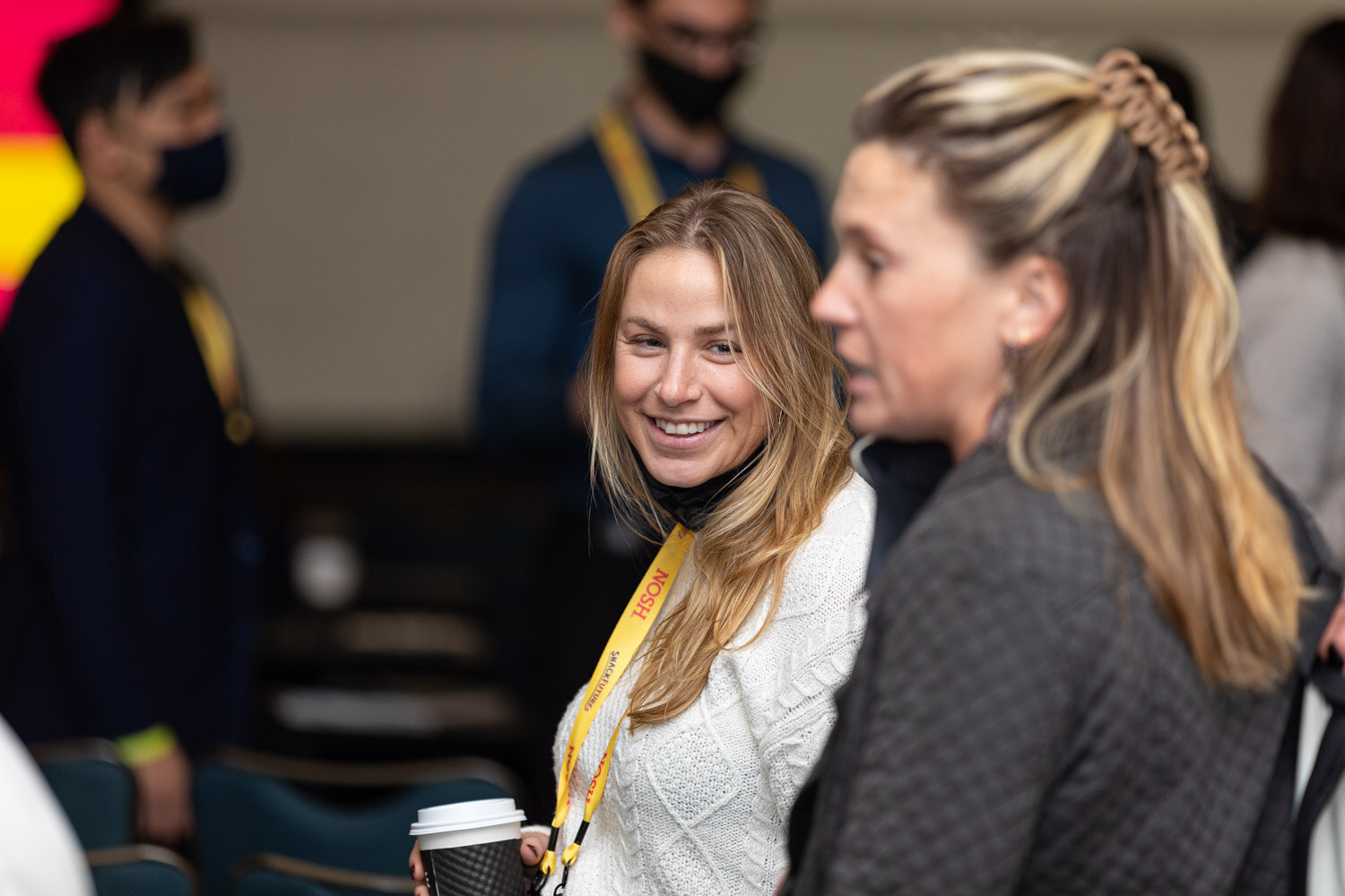 Woman with long blonde hair in a white cable-knit sweater holding a coffee cup and smiling beside a woman with a braided half-up hairstyle, both in yellow Nosh lanyards.