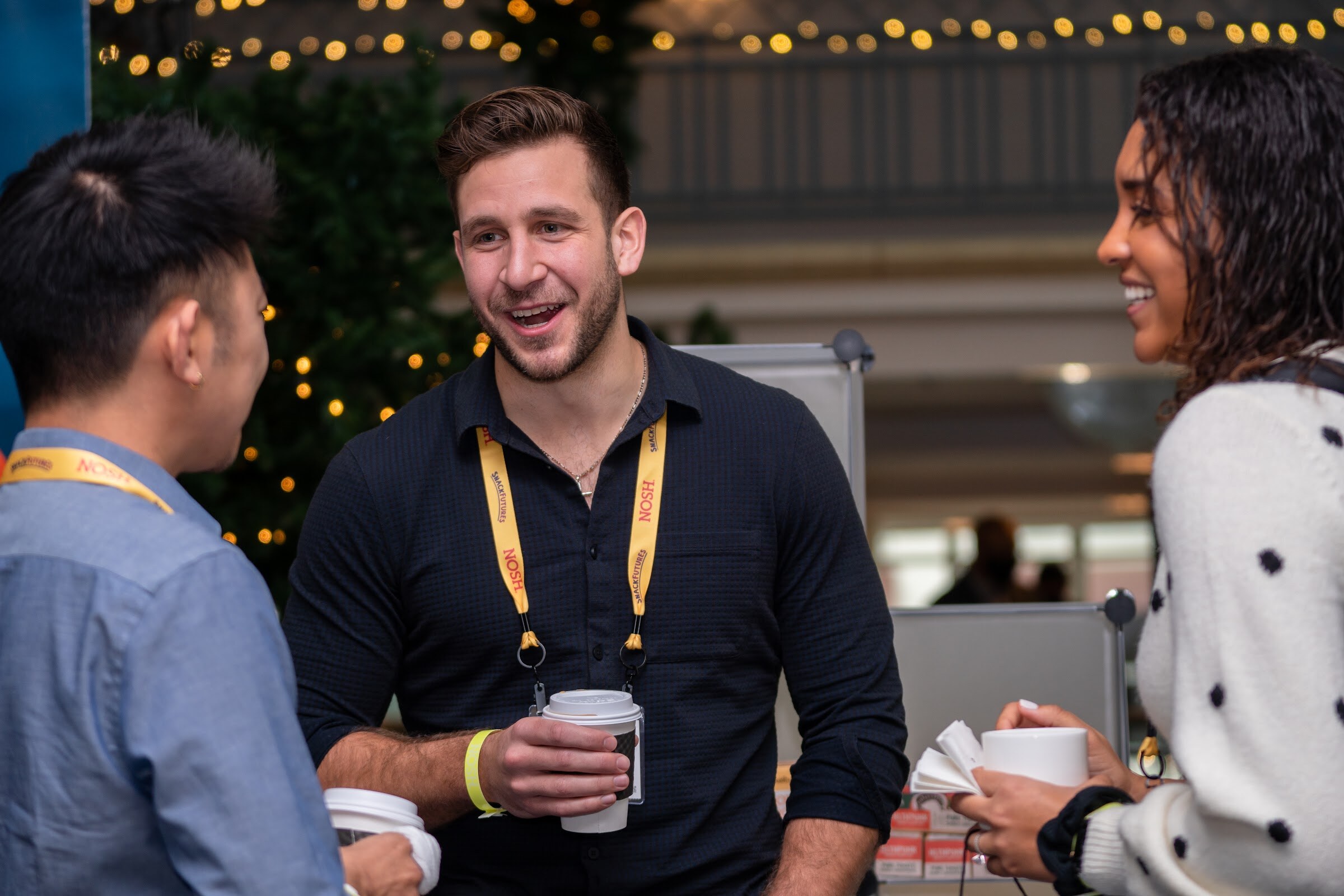 Man with dark hair and a beard in a navy shirt and yellow Nosh lanyard laughing with a woman in a cream polka-dot sweater and another attendee, with string lights and a small tree behind them.