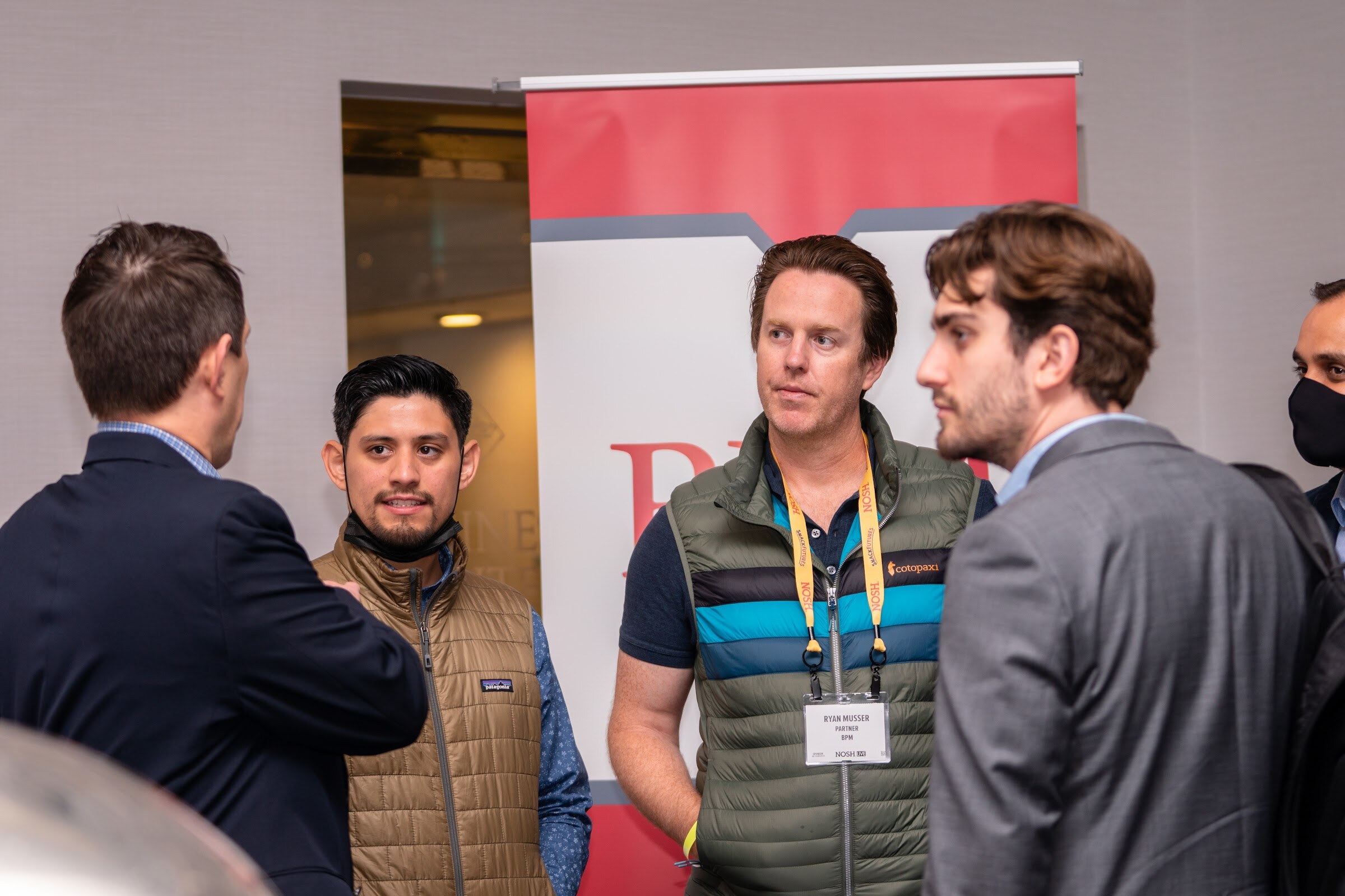 Man in a green striped puffer vest and Nosh lanyard, a man in a tan puffer vest, a man in a navy jacket and a man in a grey blazer talking in front of a red and white roll-up banner.