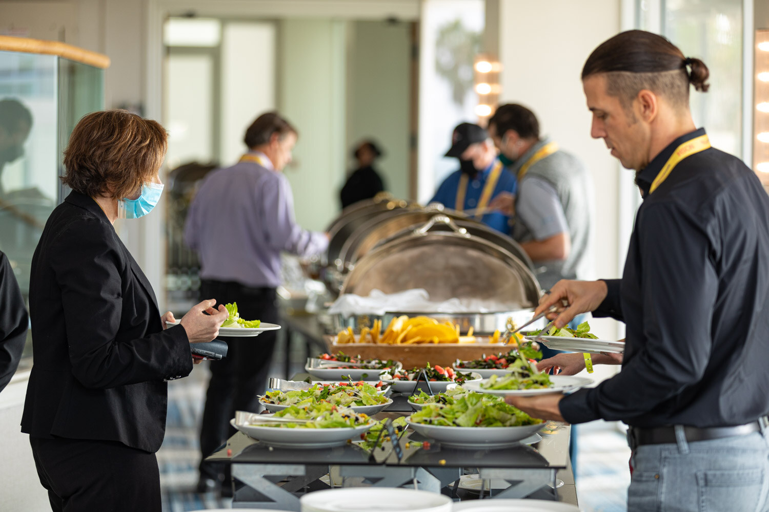 Man with a bun in a black shirt and yellow lanyard serving himself at a long buffet table with salads, while a woman in a black blazer and mask holds a plate beside him.