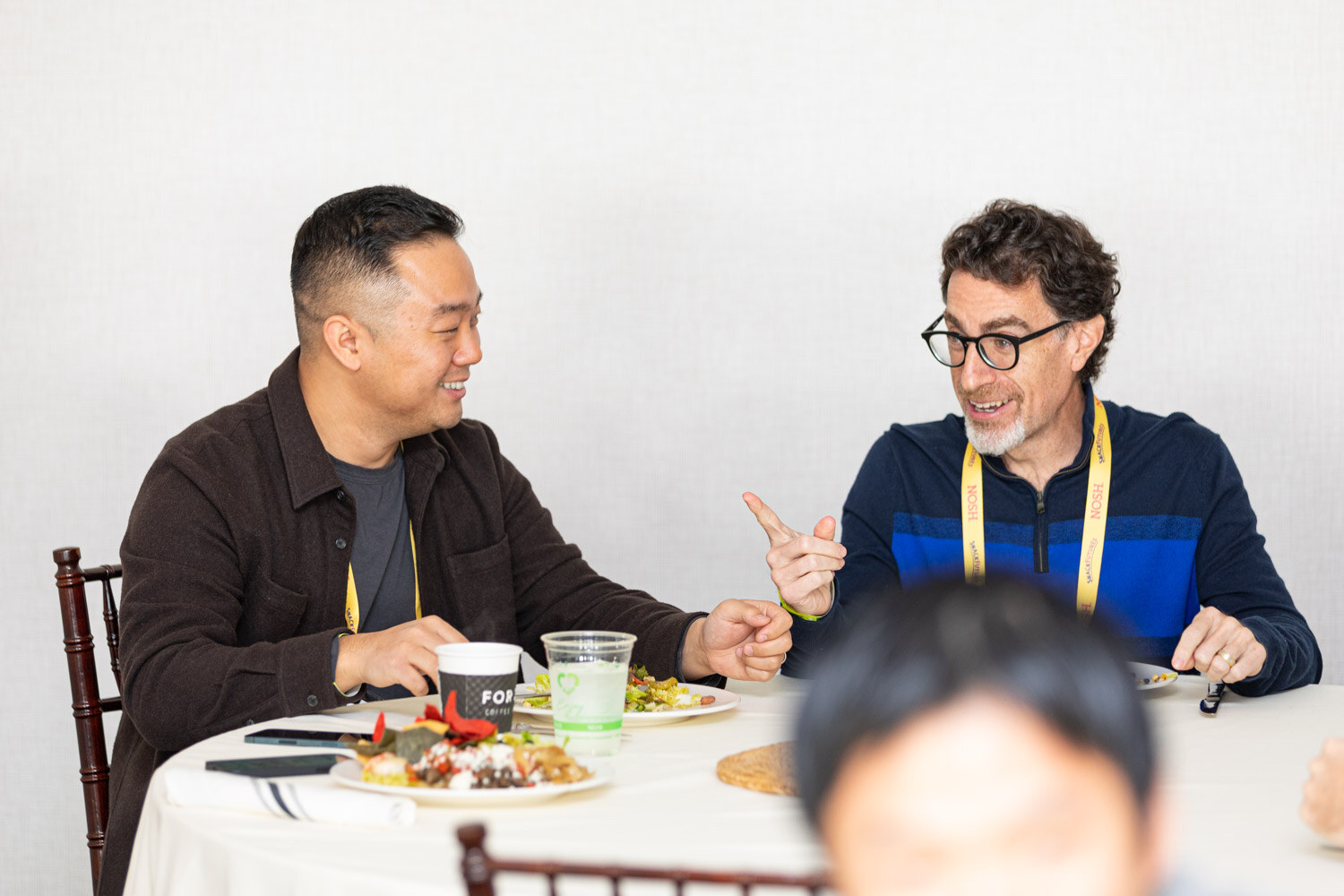 Man in a brown overshirt and man with glasses and a grey beard in a blue zip pullover, both in yellow Nosh lanyards, talking over plates of food at a round table with a white tablecloth.