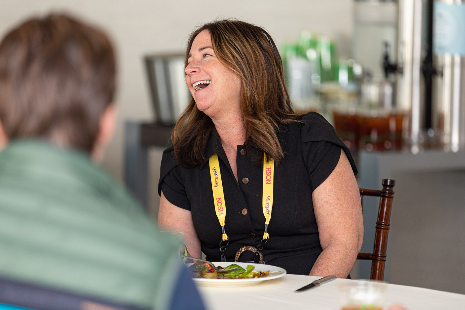 Woman with long brown hair in a black short-sleeve dress and a yellow Nosh lanyard laughing, seated at a table with a plate of salad, with a man in a green vest in the left foreground.