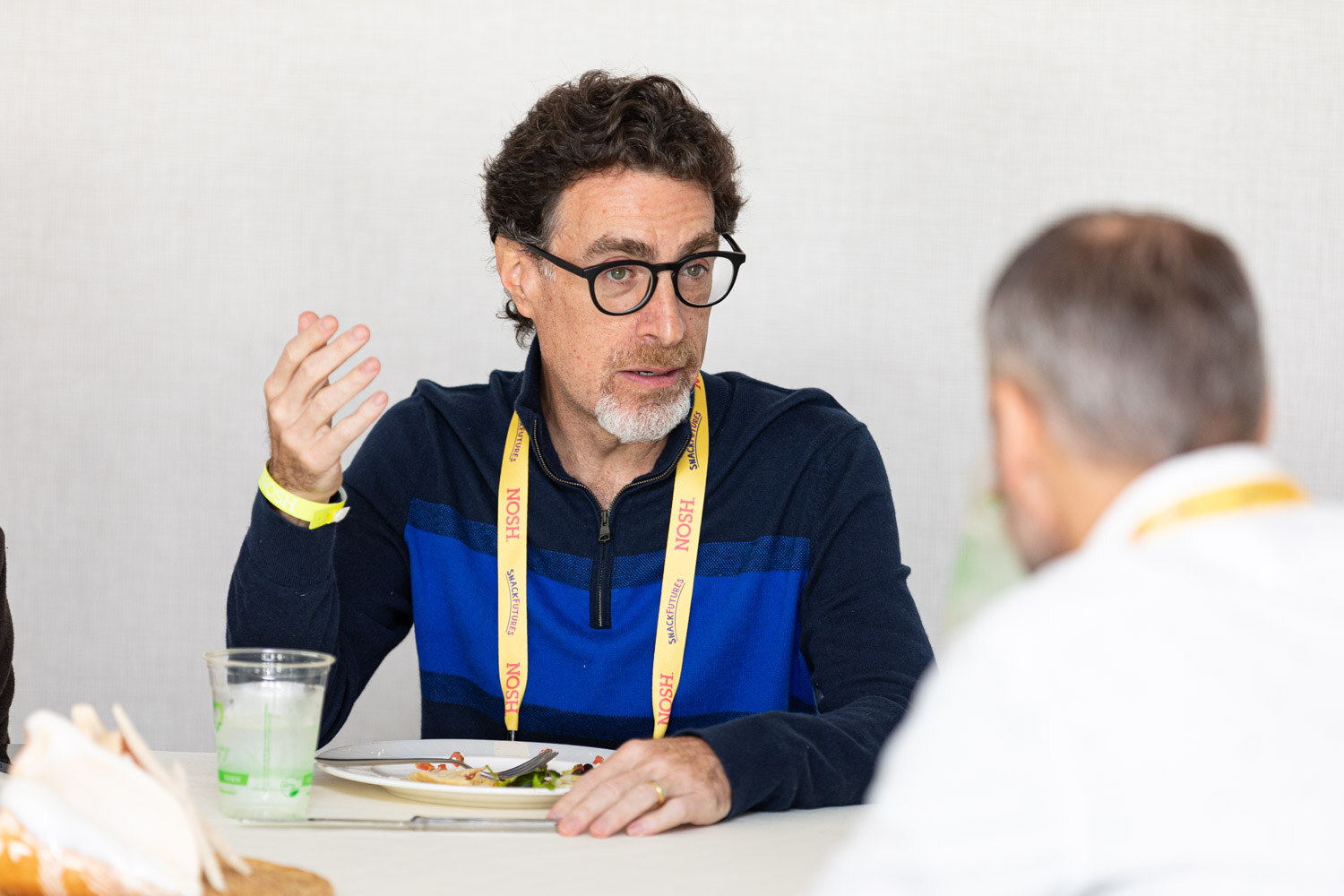 Man with dark curly hair, black glasses and a grey beard in a navy and blue zip pullover and a yellow Nosh lanyard gesturing with one hand, with a plate and a water glass in front of him.
