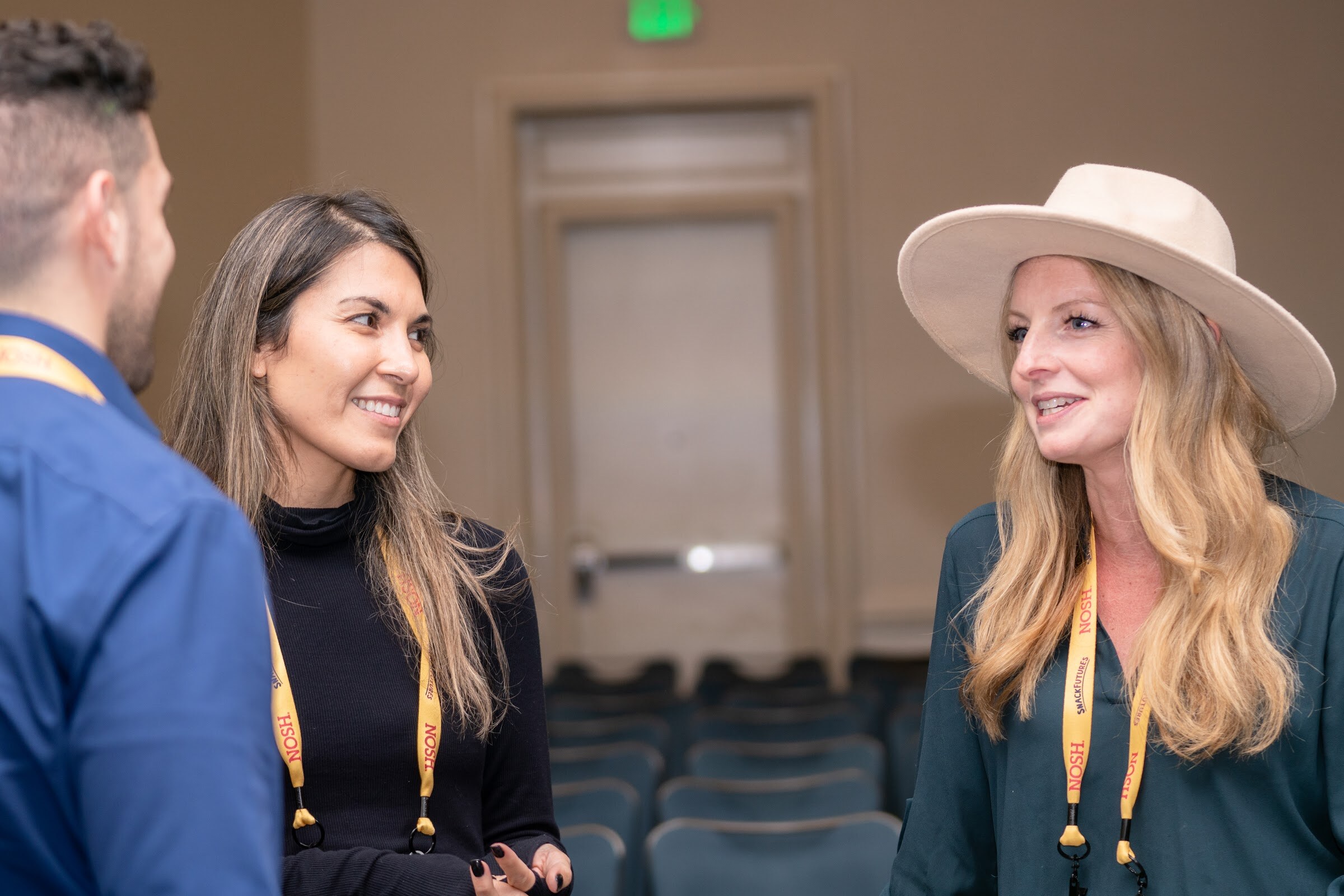 Woman with long brown hair in a black turtleneck and woman in a wide-brim cream hat and green blouse, both in yellow Nosh lanyards, smiling, with a man in a blue shirt at the left.