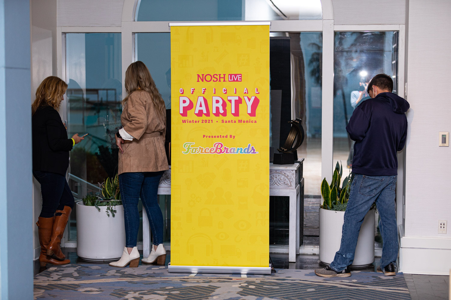 Yellow roll-up banner reading Nosh Live Official Party Winter 2021 Santa Monica Presented by ForceBrands, with three guests standing near a glass door and planters.