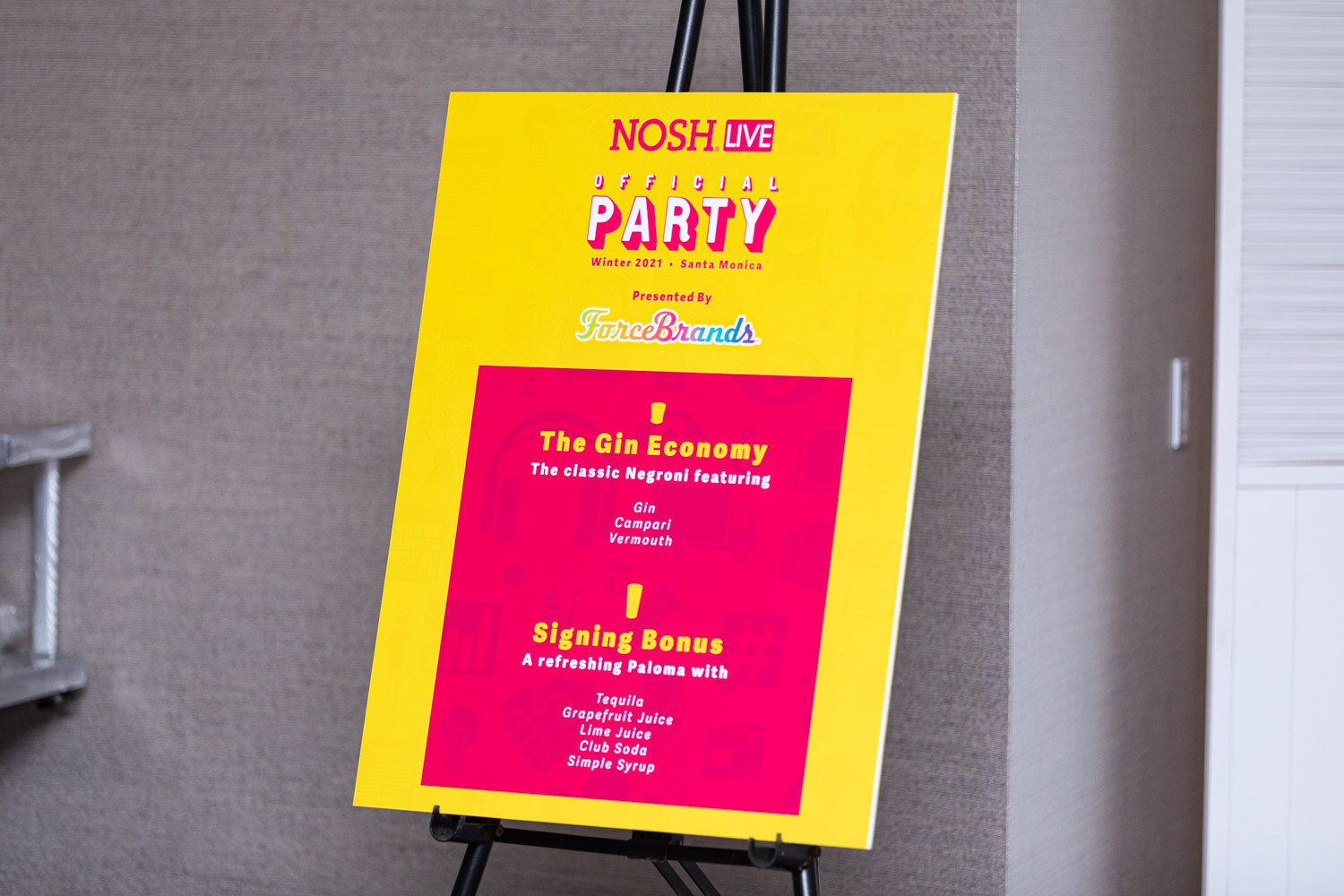 Yellow and pink easel sign reading Nosh Live Official Party Presented by ForceBrands, with The Gin Economy classic Negroni and Signing Bonus paloma recipes.