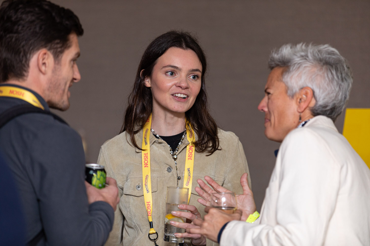 Woman with long dark hair in a tan jacket and yellow Nosh lanyard holding a drink, talking with a man holding a green can and a woman with short grey hair in a white jacket.