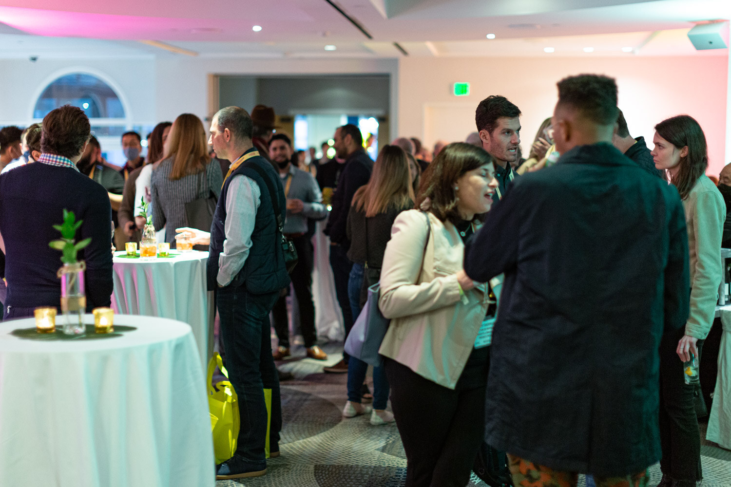 Crowded room of attendees standing and talking among cocktail tables with candles and small plants, with a yellow tote bag at the left and pink light along the ceiling.