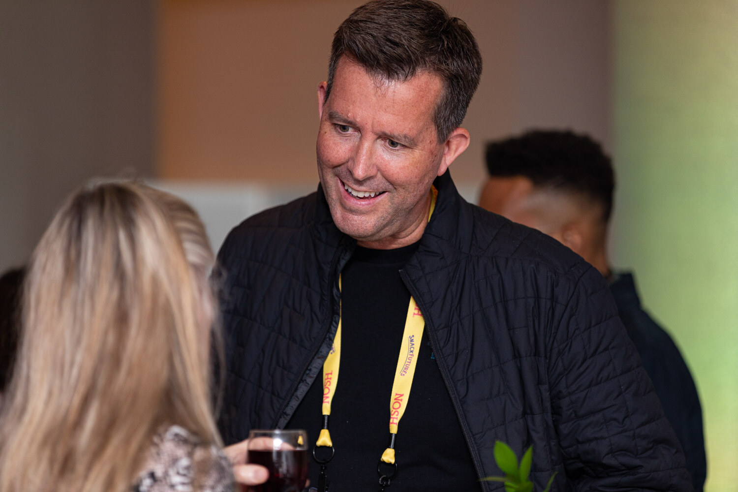 Man with short brown hair in a black quilted jacket and a yellow Nosh lanyard smiling down at a woman with long blonde hair holding a glass of wine.