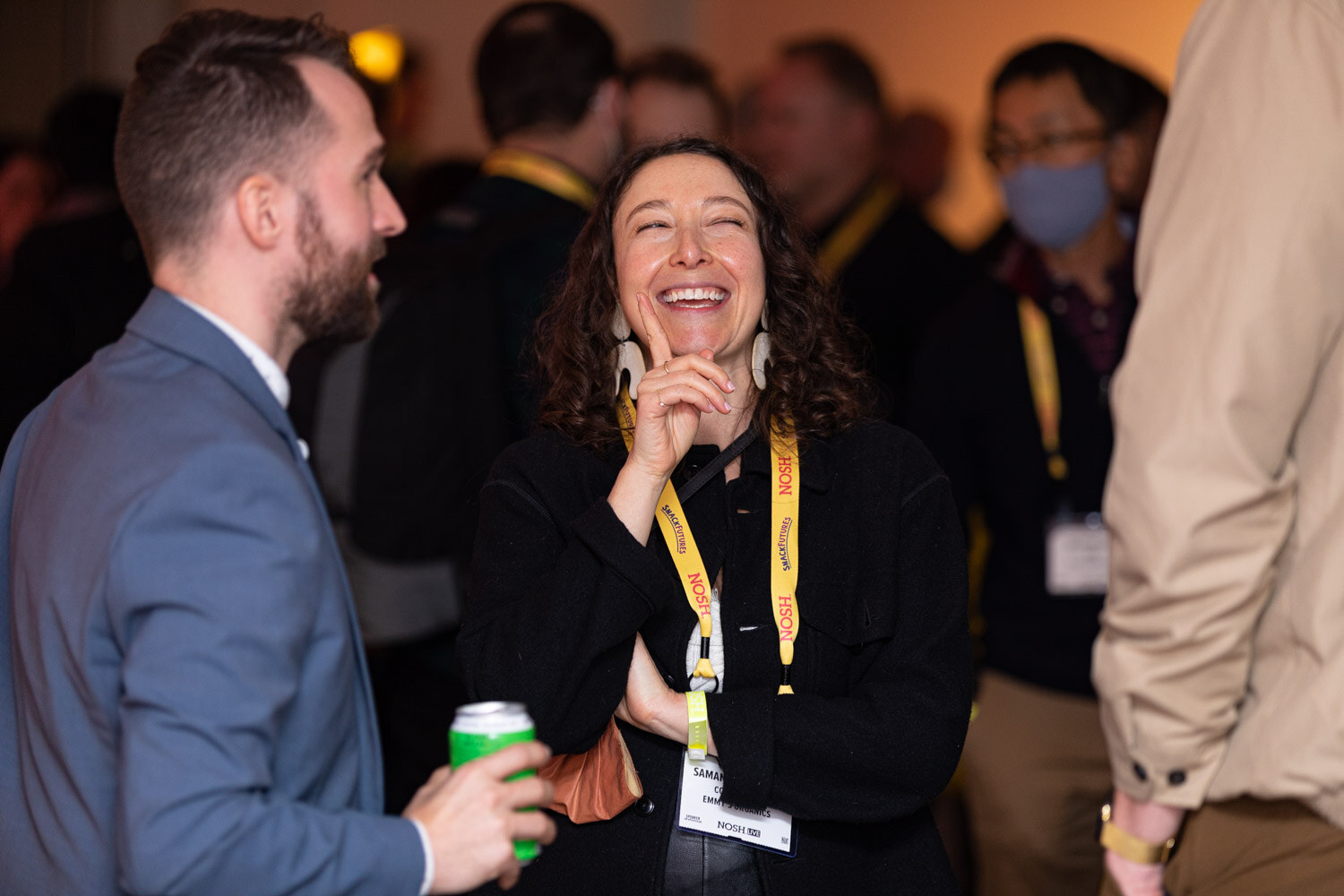 Woman with curly dark hair in a black top and yellow Nosh lanyard laughing, with a bearded man in a blue blazer holding a green can facing her.