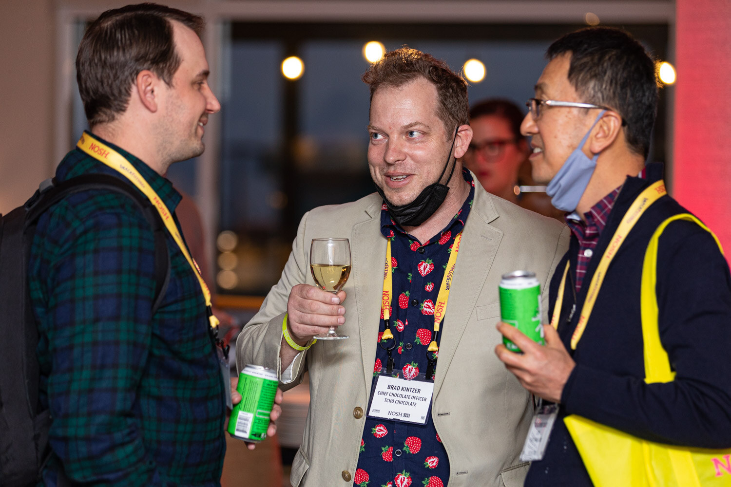 Man in a tan blazer and strawberry-print shirt holding a glass of white wine beside a man in a plaid shirt and a man in a mask with a yellow tote bag, all in Nosh lanyards.