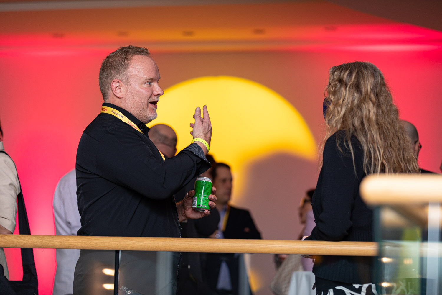 Man in a black shirt holding a green can and gesturing while talking to a woman with long wavy blonde hair, with a pink and yellow lit wall behind a wooden railing.