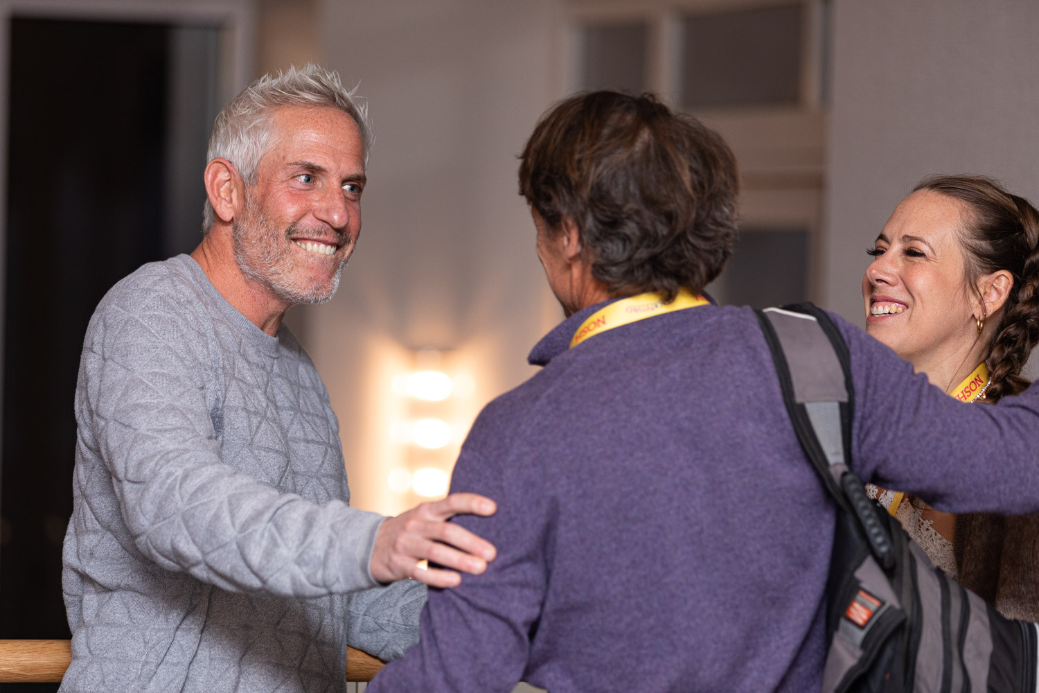 Man with spiky grey hair and a grey quilted sweater smiling and reaching toward a man with a backpack in a purple fleece, with a woman with a braid laughing at the right.