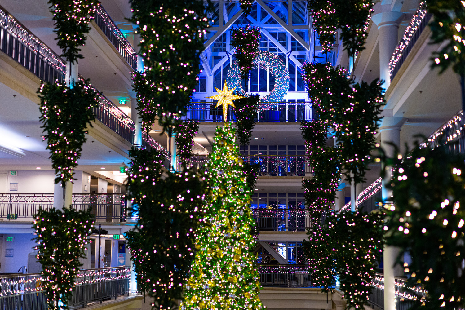 Multi-story atrium with a tall lit Christmas tree, hanging evergreen garlands with white lights, a gold star and a lighted wreath under a skylight.