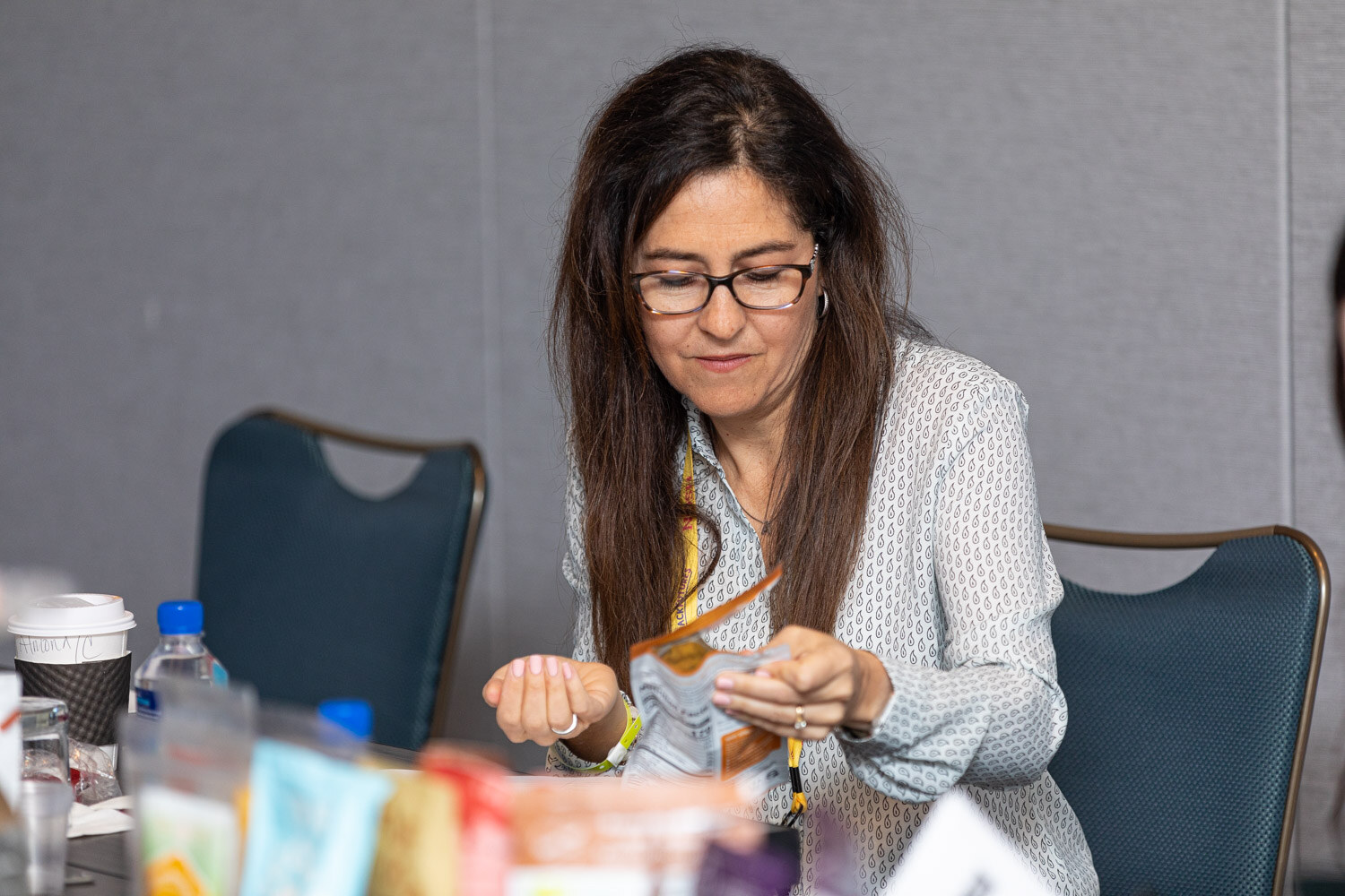 Woman with glasses and long dark hair opening a small foil package at a table crowded with product samples and a coffee cup.