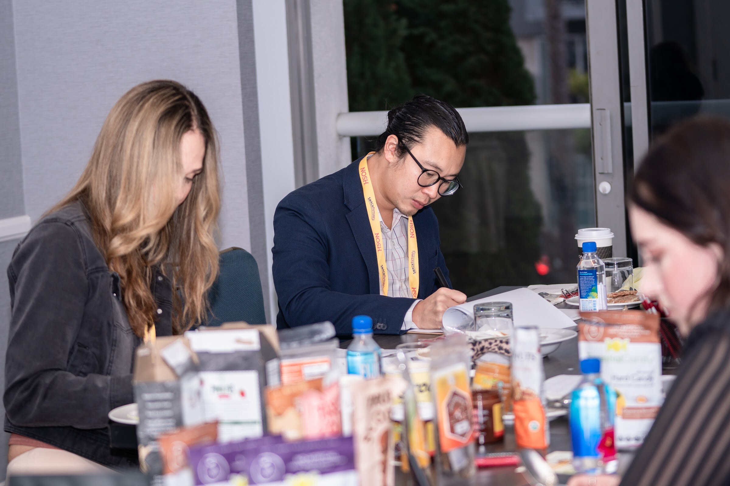 Two judges at a table filled with packaged snack samples, water bottles and paper score sheets, one writing and one with her head down.