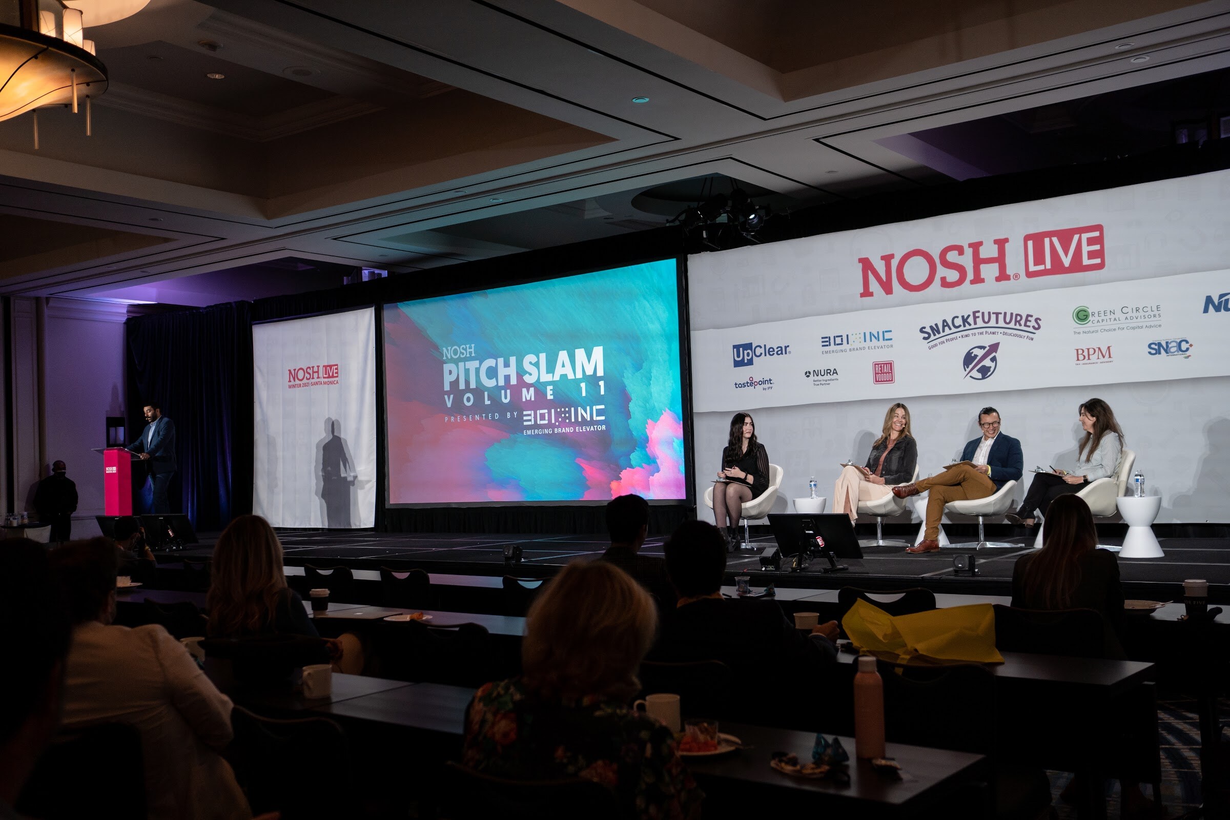 Wide shot of a ballroom stage with a large screen reading Pitch Slam Volume 11, a Nosh Live sponsor backdrop and four judges seated in white chairs.