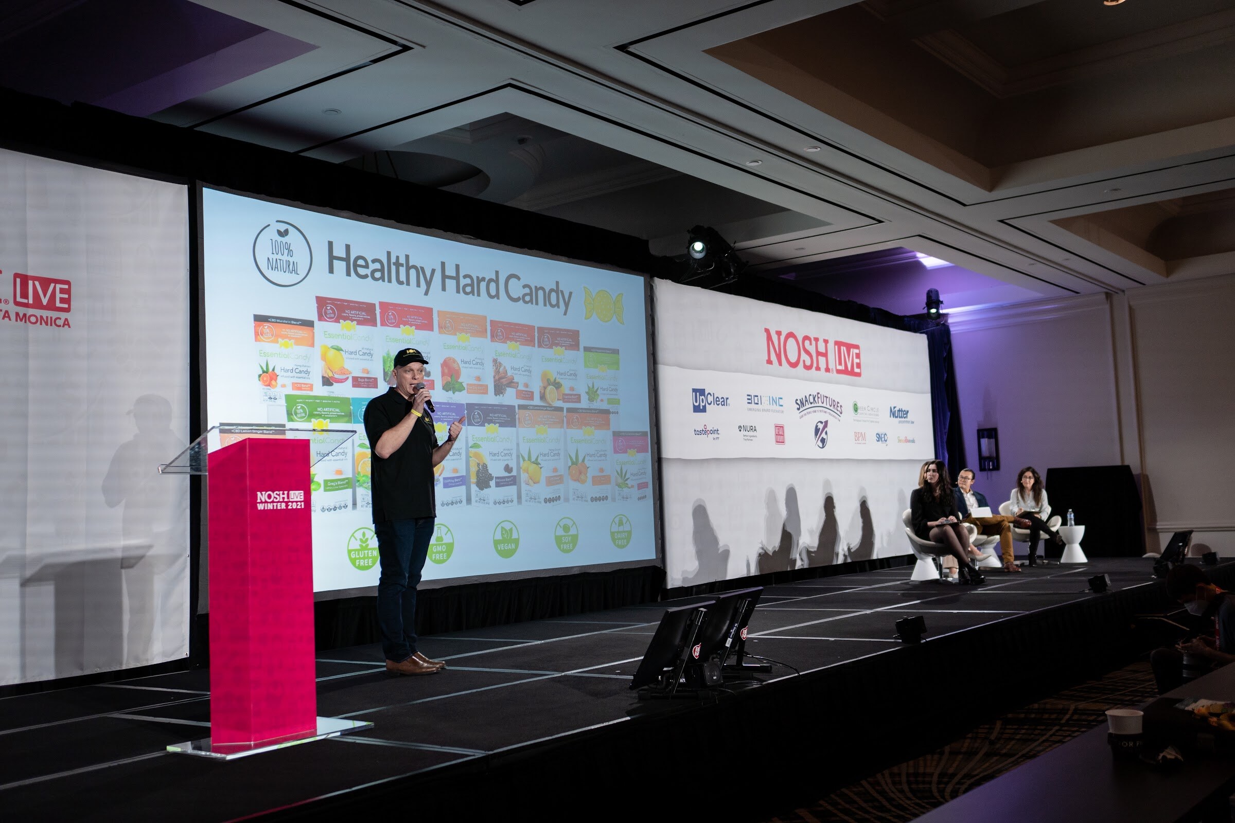 Man in a black polo and cap speaking at a pink podium in front of a screen that reads Healthy Hard Candy with rows of candy packages.