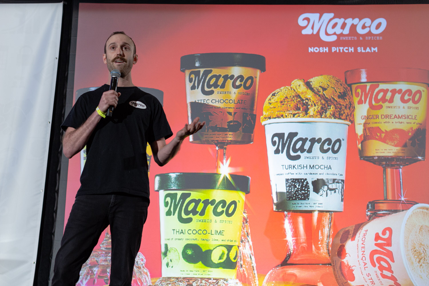 Man in a black Marco t-shirt holding a microphone in front of a red screen showing Marco Sweets and Spices ice cream pints and the words Nosh Pitch Slam.