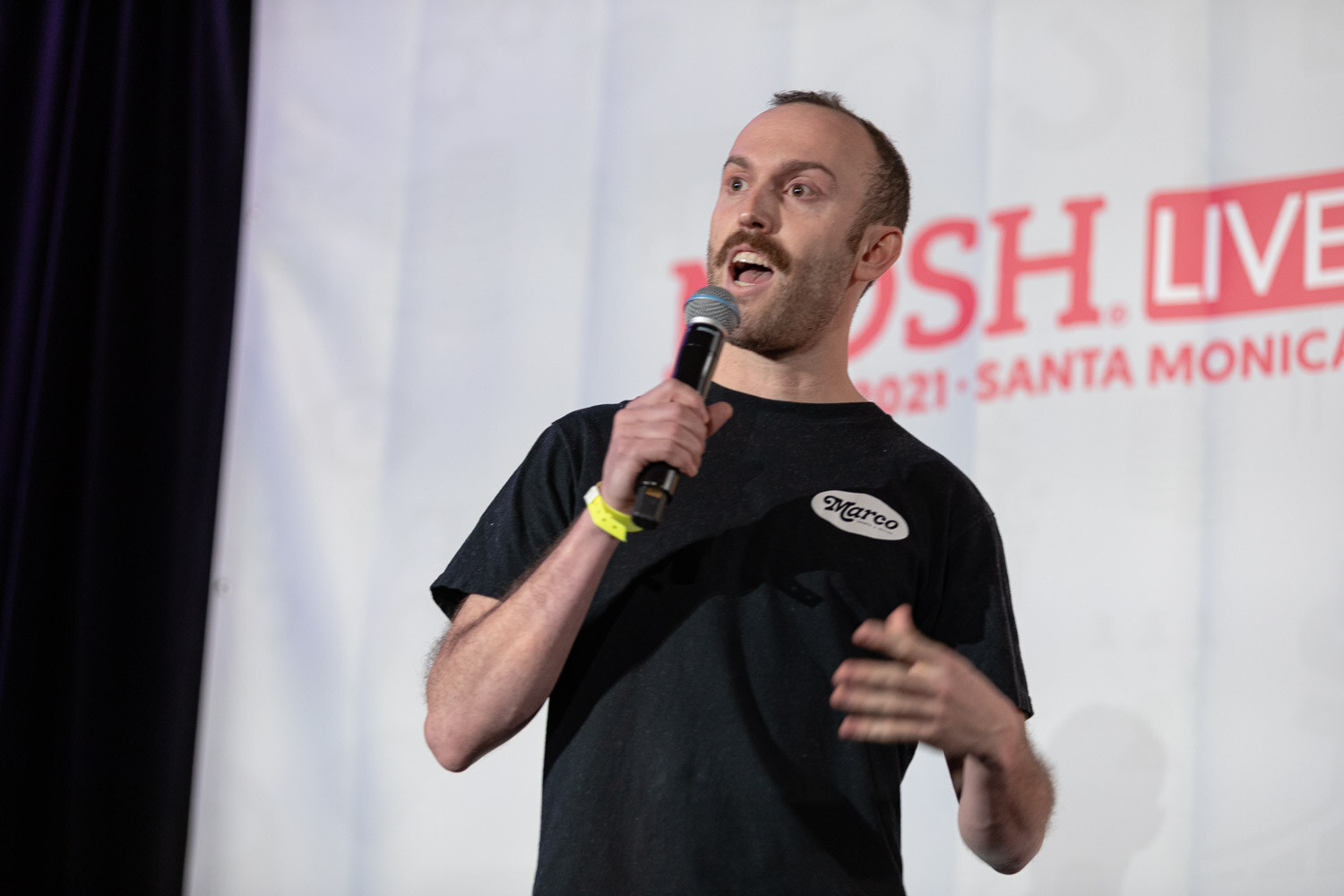 Man in a black t-shirt with a Marco logo patch speaking into a microphone in front of a white Nosh Live Winter 2021 Santa Monica banner.