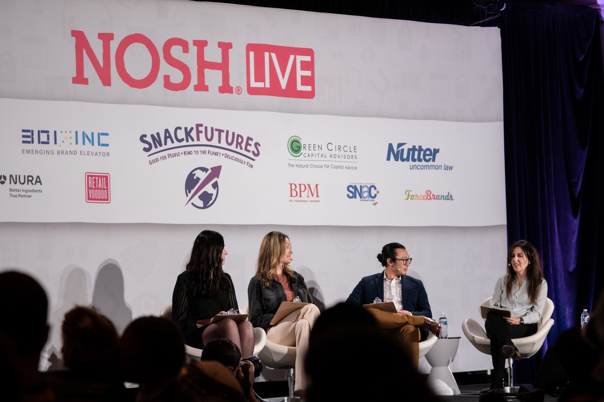 Four people seated on white chairs on a stage in front of a Nosh Live banner with sponsor logos including 301 Inc, SnackFutures, Green Circle Capital Advisors, Nutter, BPM and SNAC.