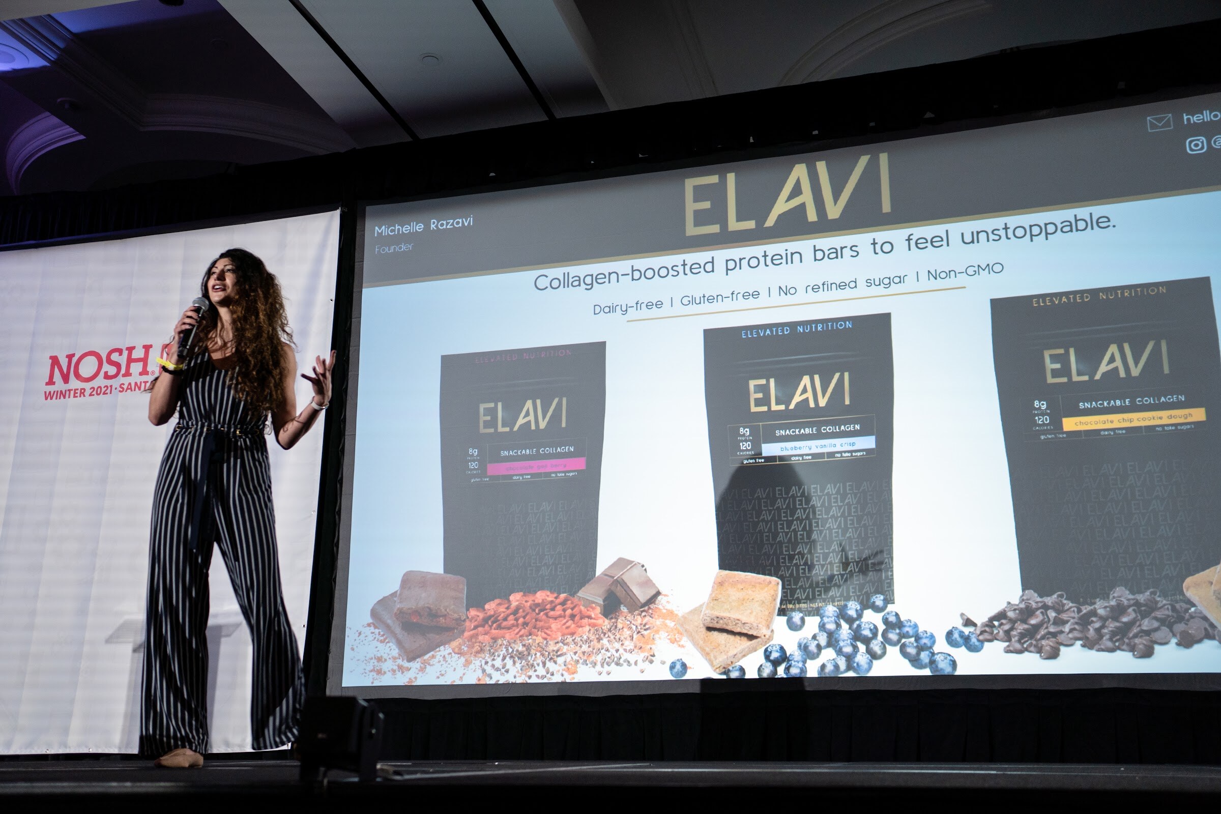 Woman with long curly hair in a navy pinstriped jumpsuit holding a microphone on stage beside a large screen showing Elavi collagen-boosted protein bars.