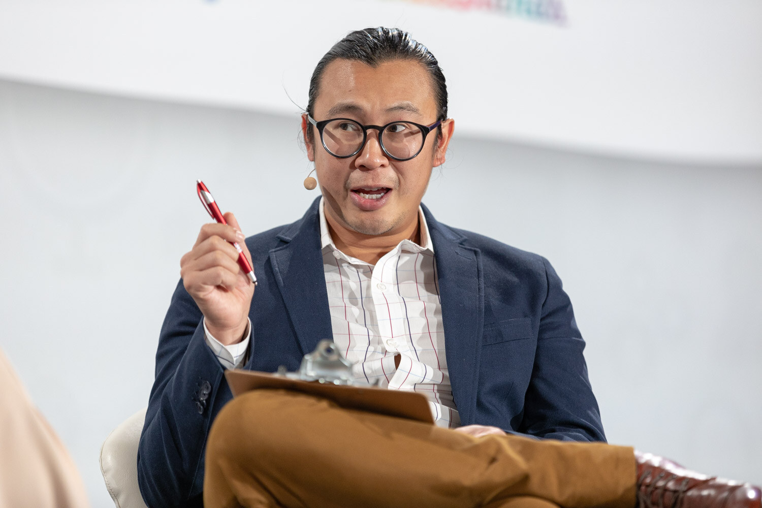 Man with glasses and a bun in a navy blazer holding a red pen in one hand and a clipboard on his lap.