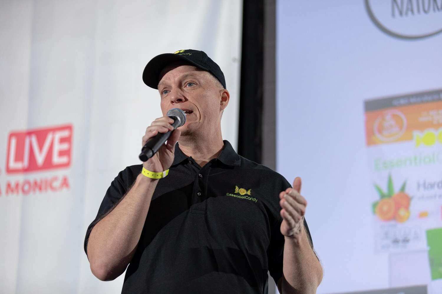 Man in a black polo shirt with an EssentialCandy logo and a black cap holding a microphone, with part of a Nosh Live banner and slide behind him.