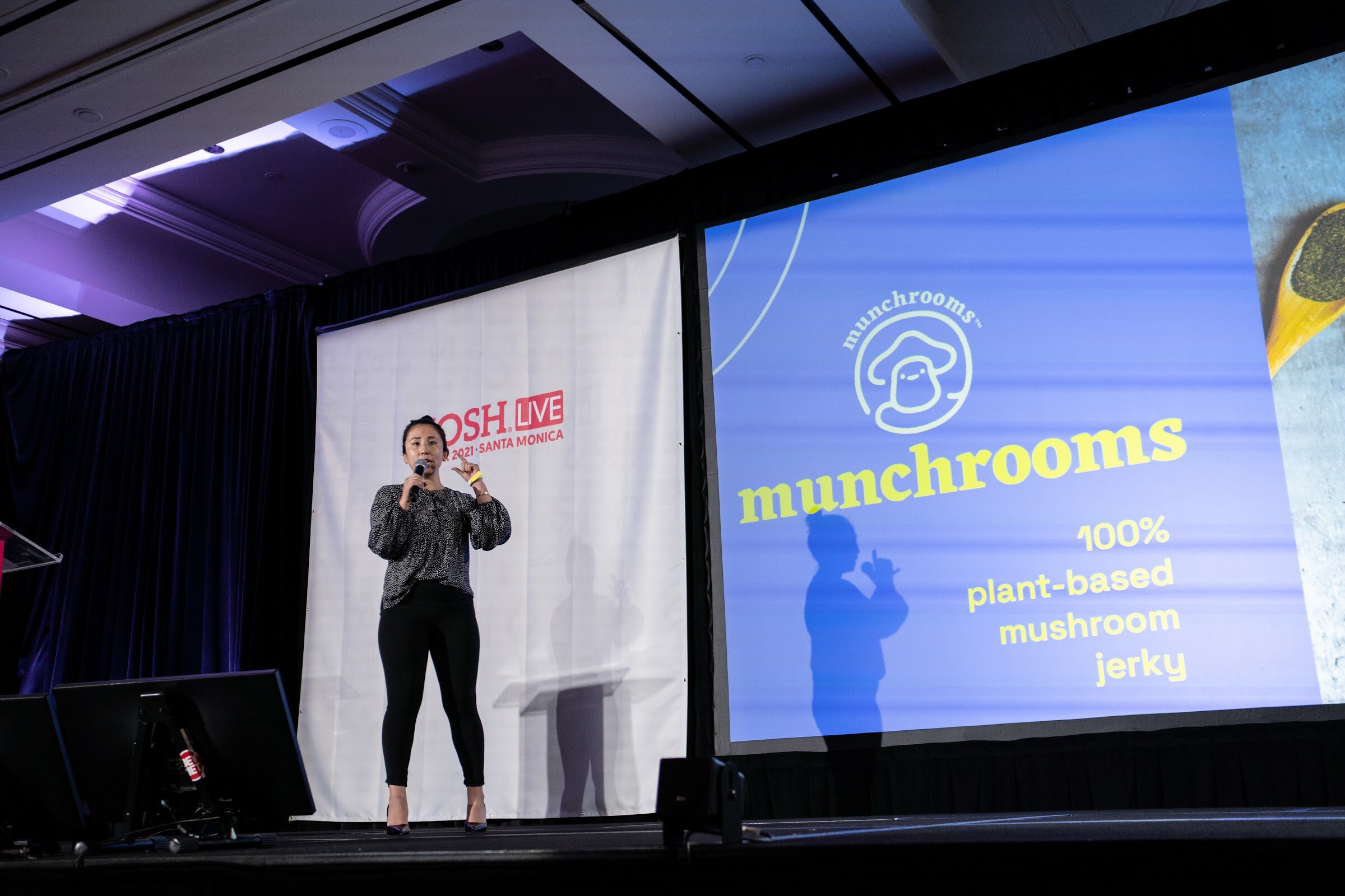 Woman in a patterned top and black pants on a dark stage beside a large blue screen reading munchrooms 100% plant-based mushroom jerky.
