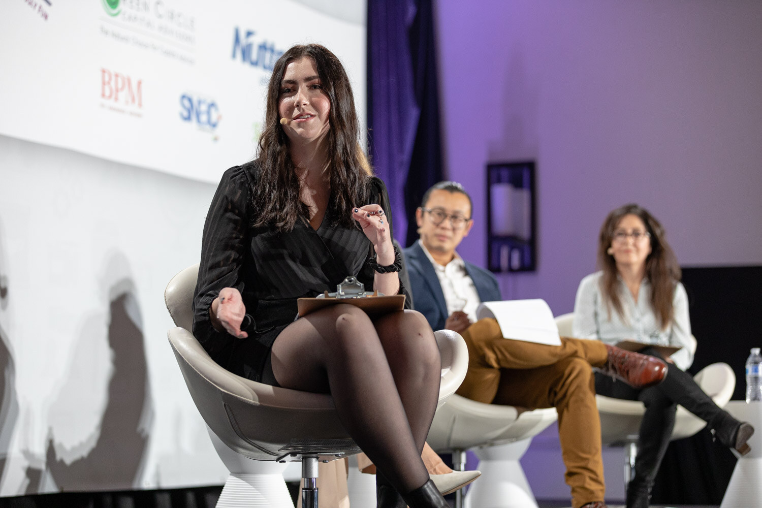 Woman with long dark hair in a black dress seated in a white chair with a clipboard, with two other judges seated behind her on a stage.
