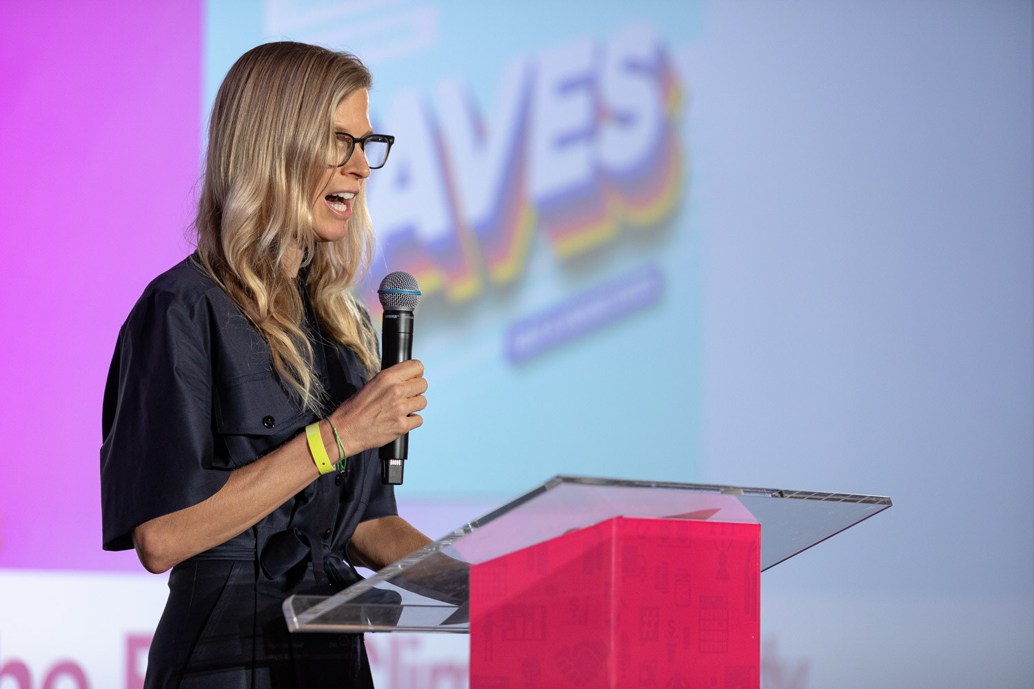 Woman with long blonde hair and dark glasses in a navy dress speaking into a microphone at a clear podium with a pink base, in front of a bright slide reading Faves.