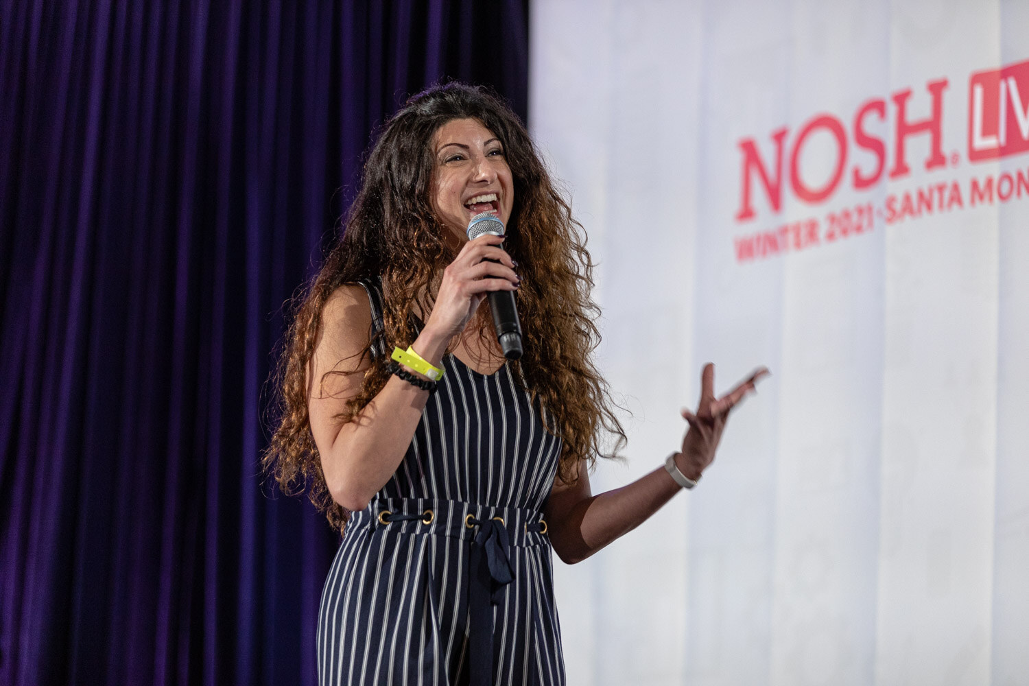 Woman with long curly hair in a navy striped jumpsuit talking into a handheld microphone in front of a white Nosh Live Winter 2021 Santa Monica banner.