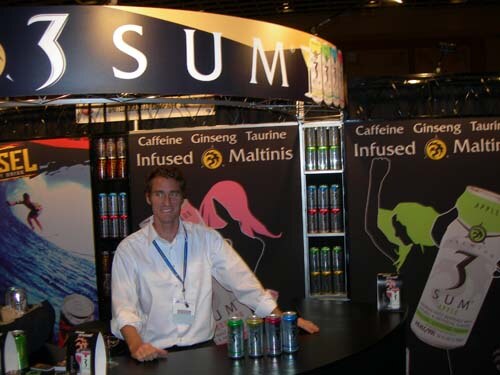 A smiling man in a white shirt and lanyard stands behind a curved counter with 3 Sum cans, in front of signs reading Caffeine Ginseng Taurine Infused Maltinis.