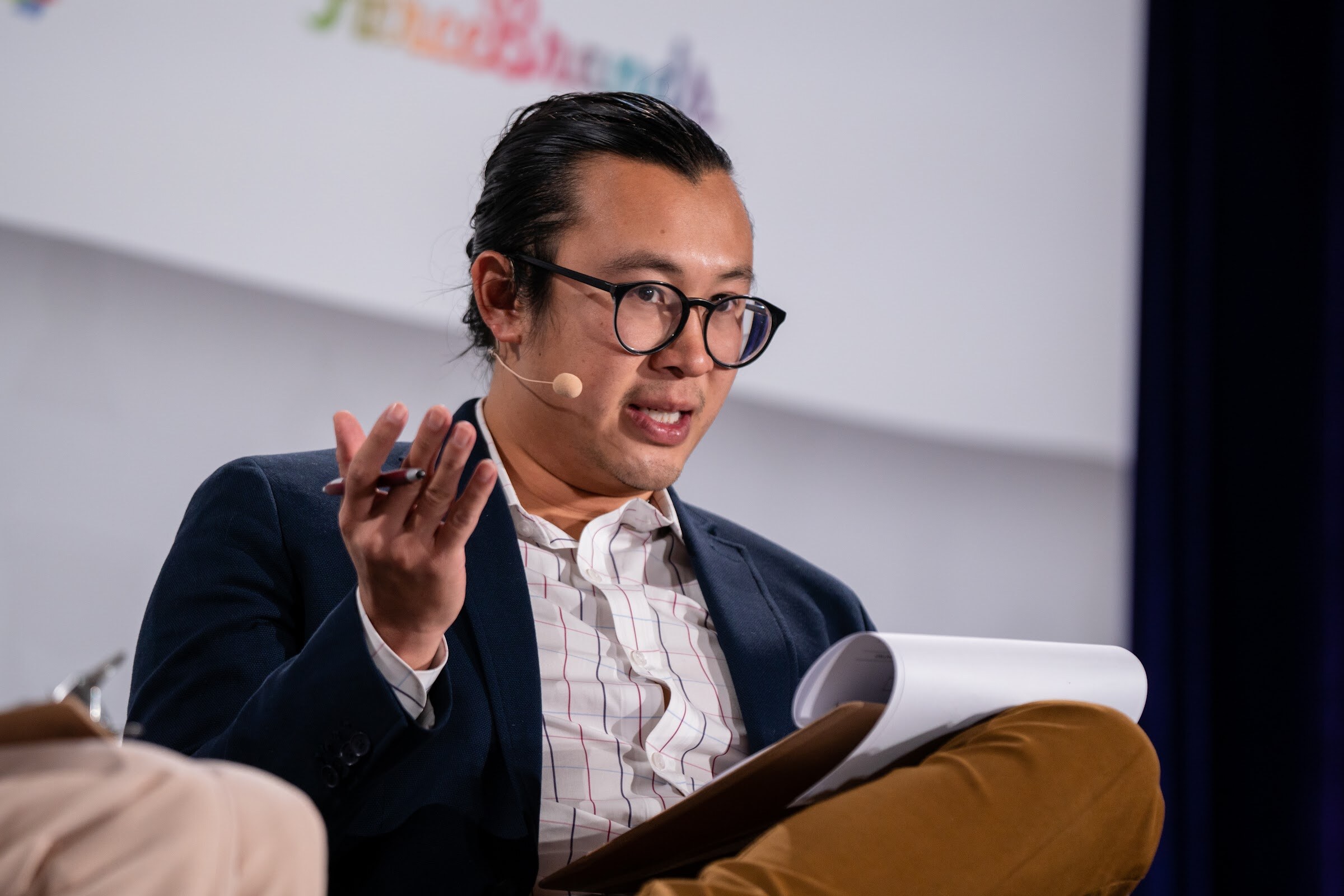 Man with glasses and a bun wearing a navy blazer and a headset microphone, gesturing with a pen above a notepad on his knee.