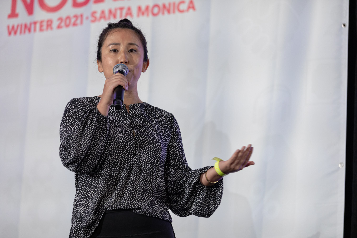 Close view of a woman in a black and white polka-dot blouse holding a microphone in front of a white Nosh Live banner.