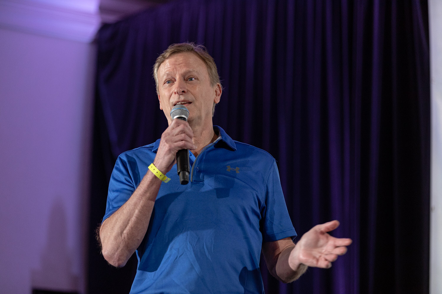 Man in a bright blue polo holding a microphone and gesturing in front of a dark purple curtain.