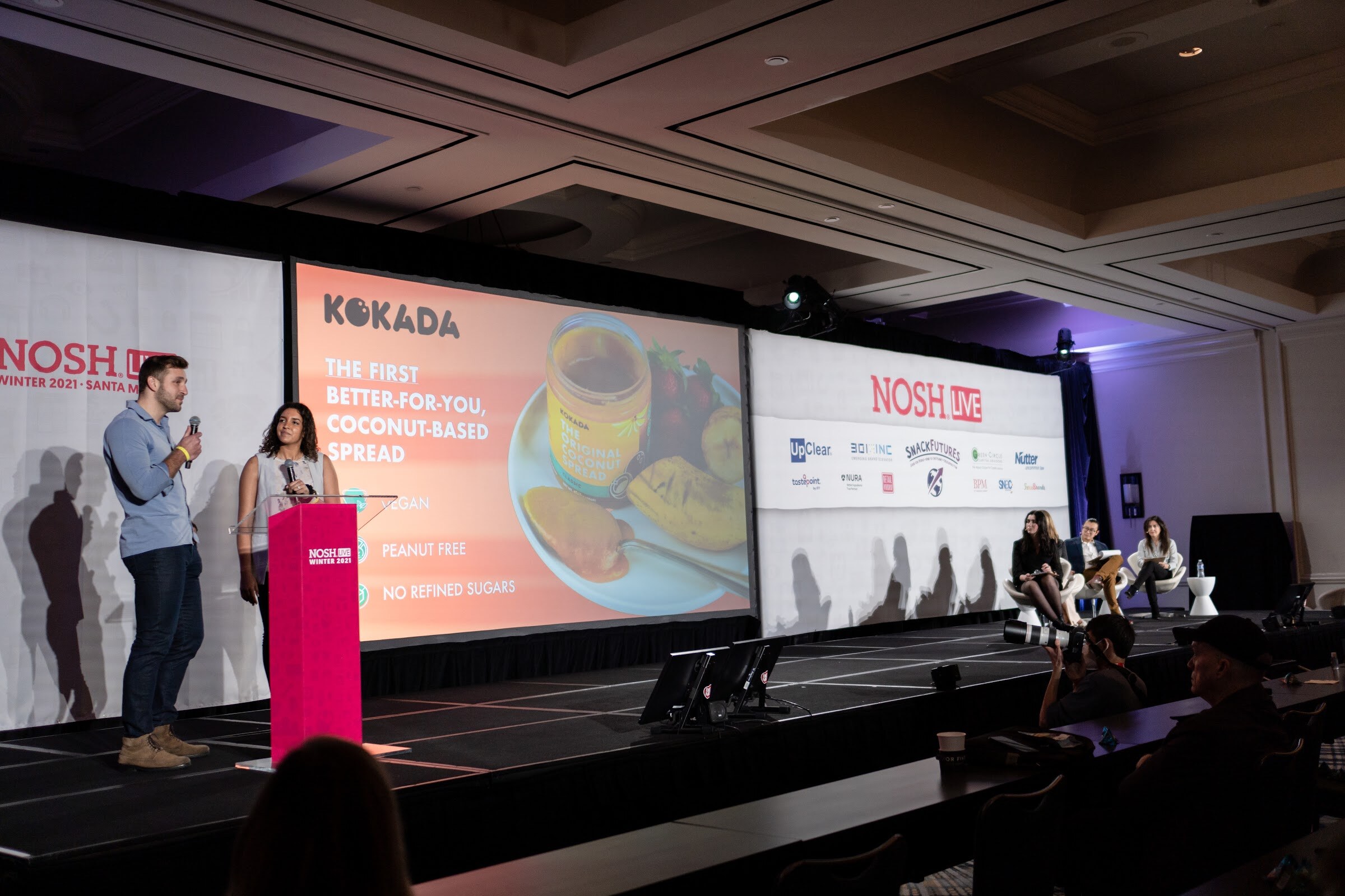 Man and woman at a pink podium beside a large screen showing a Kokada slide about a vegan, peanut-free coconut spread, with judges seated at the far right.