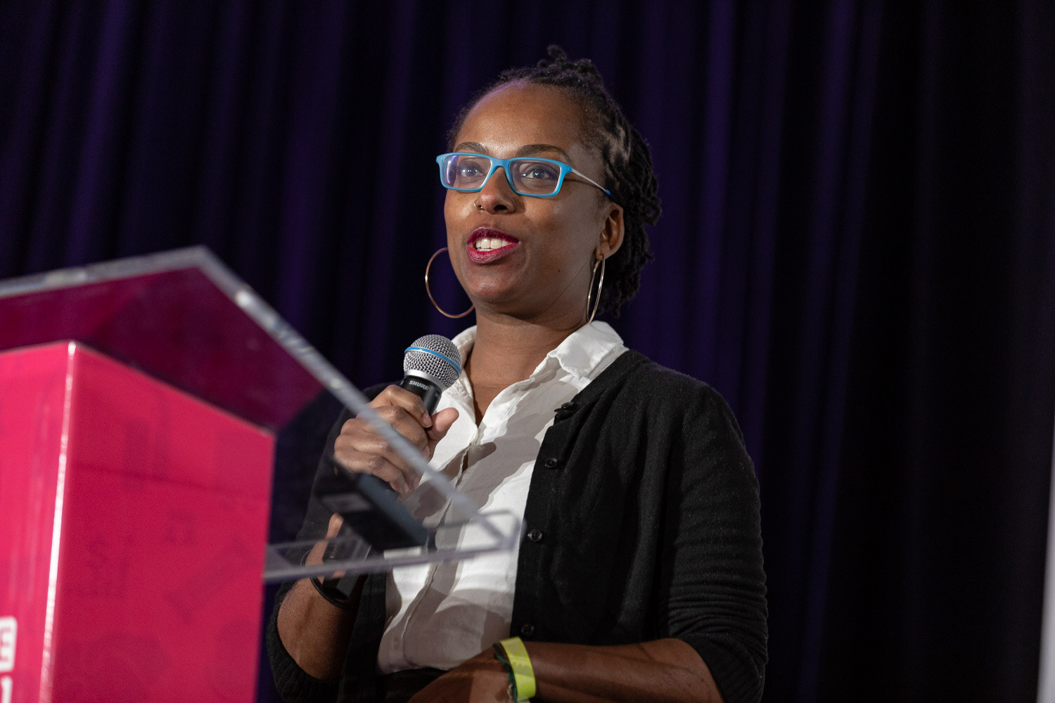 Woman with blue glasses, hoop earrings, a white collared shirt and black cardigan holding a microphone beside a pink podium against a dark curtain.