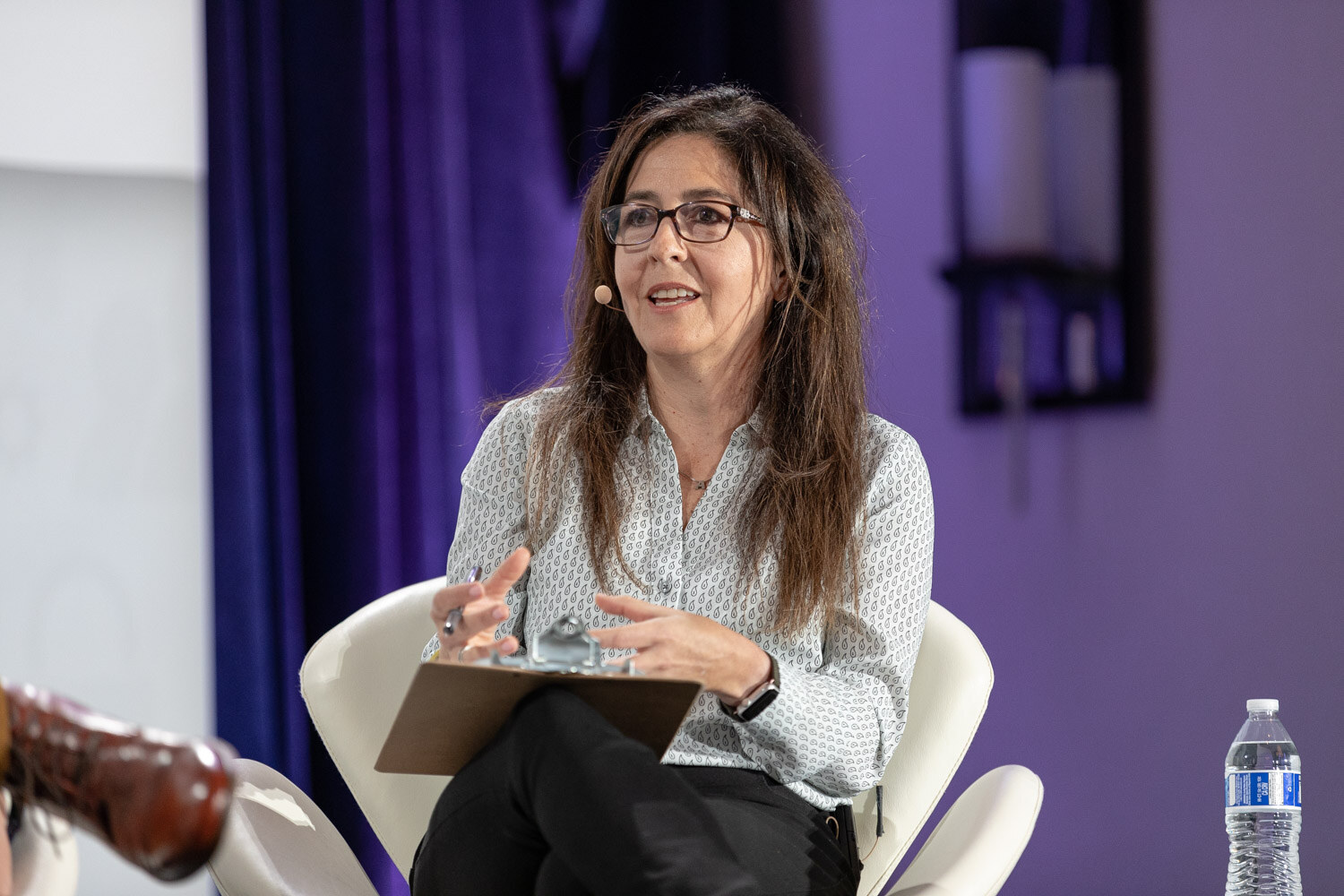 Woman with glasses and long brown hair in a light patterned blouse sitting in a white chair with a clipboard on her lap in front of a purple curtain.