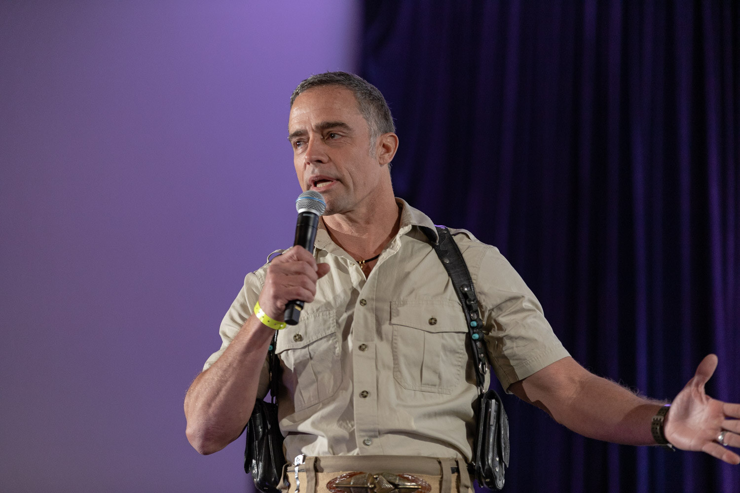 Man with short grey hair in a khaki shirt and shoulder harness holding a microphone with one arm outstretched in front of a purple curtain.