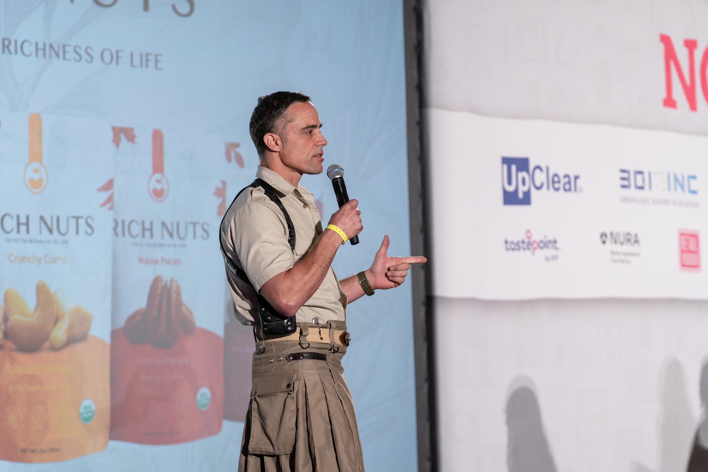 Man in a khaki shirt with shoulder straps and a khaki kilt holding a microphone beside a screen showing Rich Nuts packages.