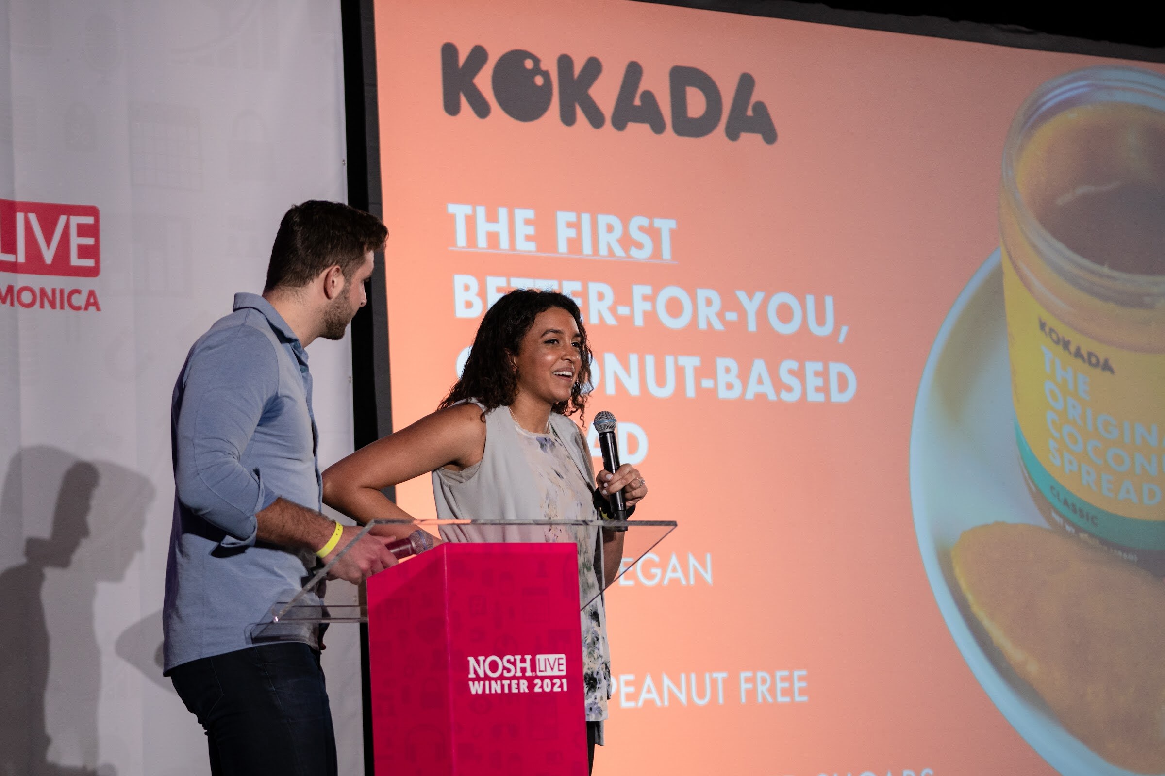 Man in a blue shirt and woman in a grey top at a clear podium with a pink base, in front of a salmon-colored Kokada slide.