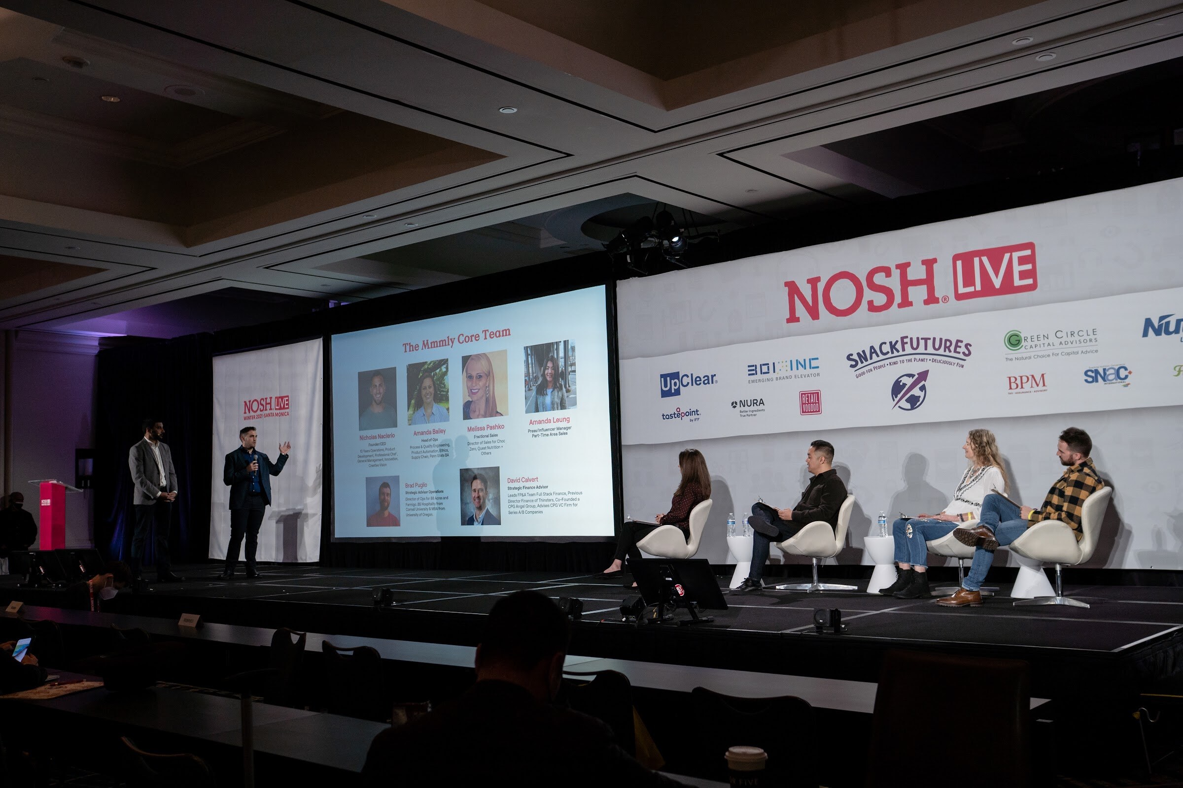 Wide view of the Nosh Live stage with a presenter and a host at the left, a slide titled The Mmmly Core Team, and four judges seated on white chairs at the right.