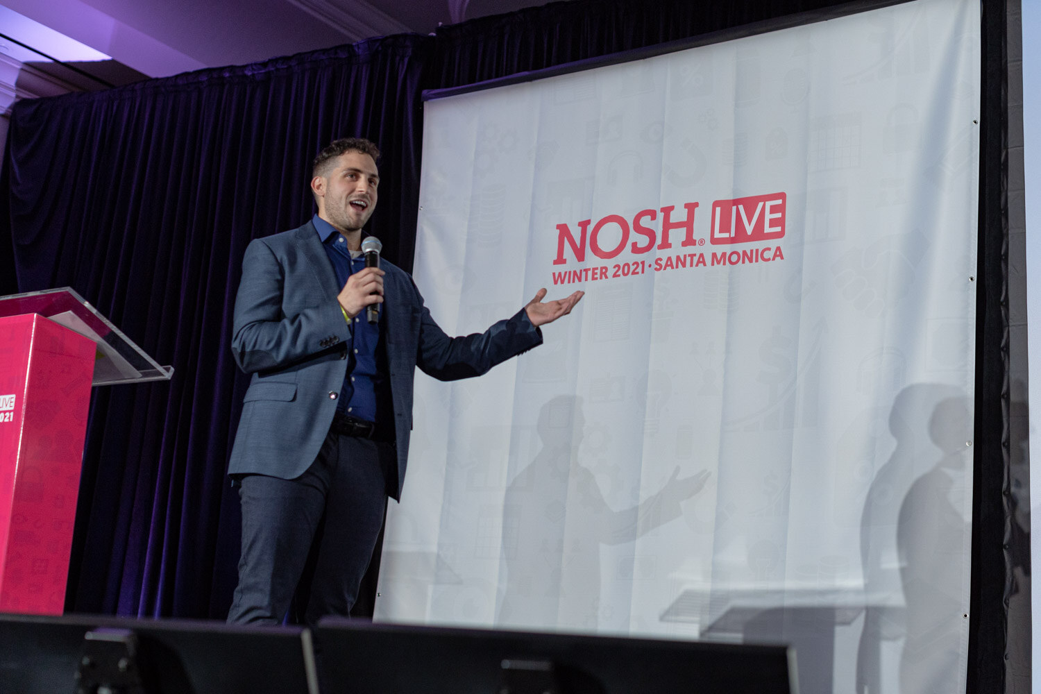 Man in a blue blazer and navy shirt holding a microphone and gesturing with an open hand in front of a white Nosh Live Winter 2021 Santa Monica banner.