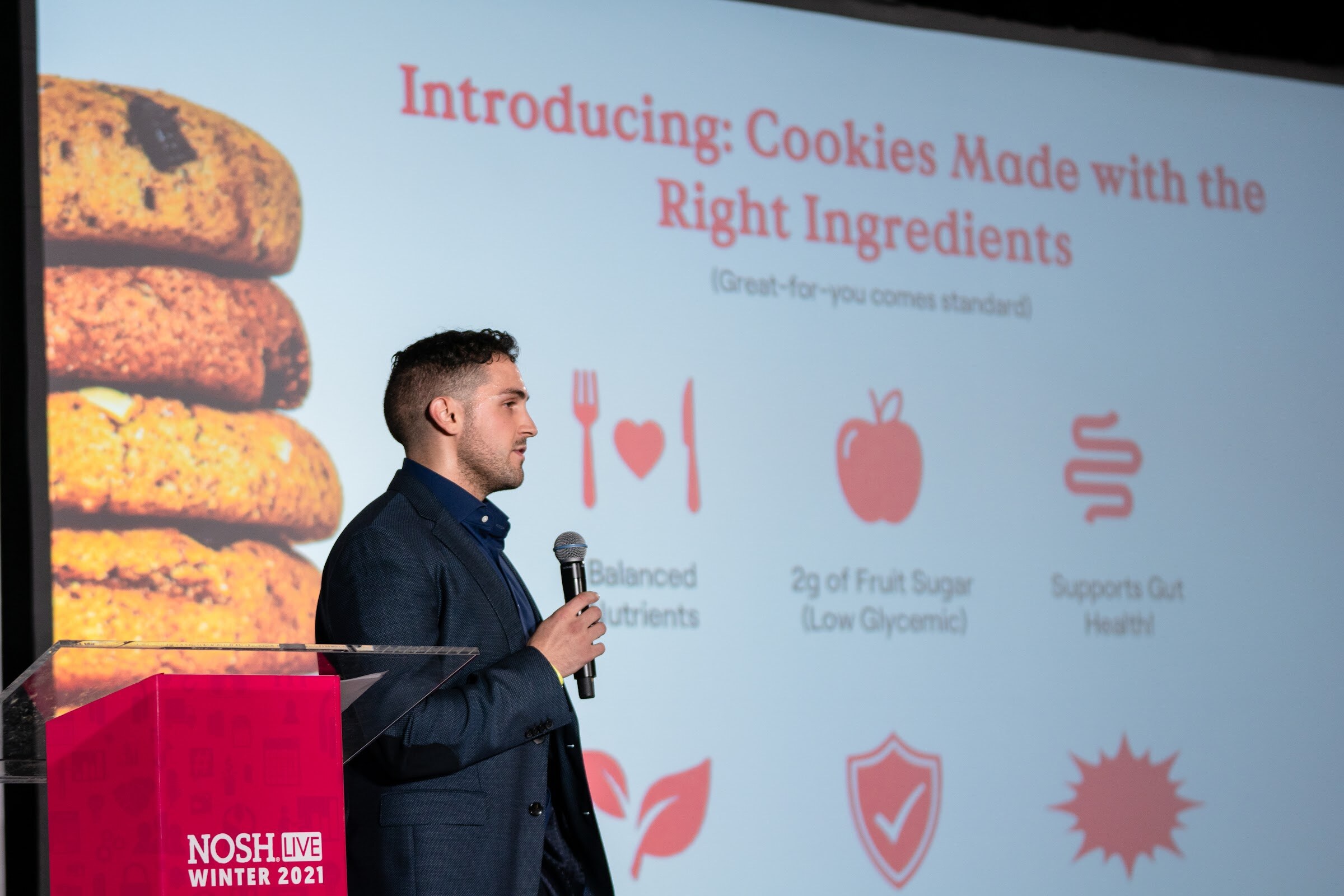 Man in a dark blazer holding a microphone at a pink Nosh Live podium in front of a slide about cookies made with the right ingredients, with icons for balanced nutrients and fruit sugar.