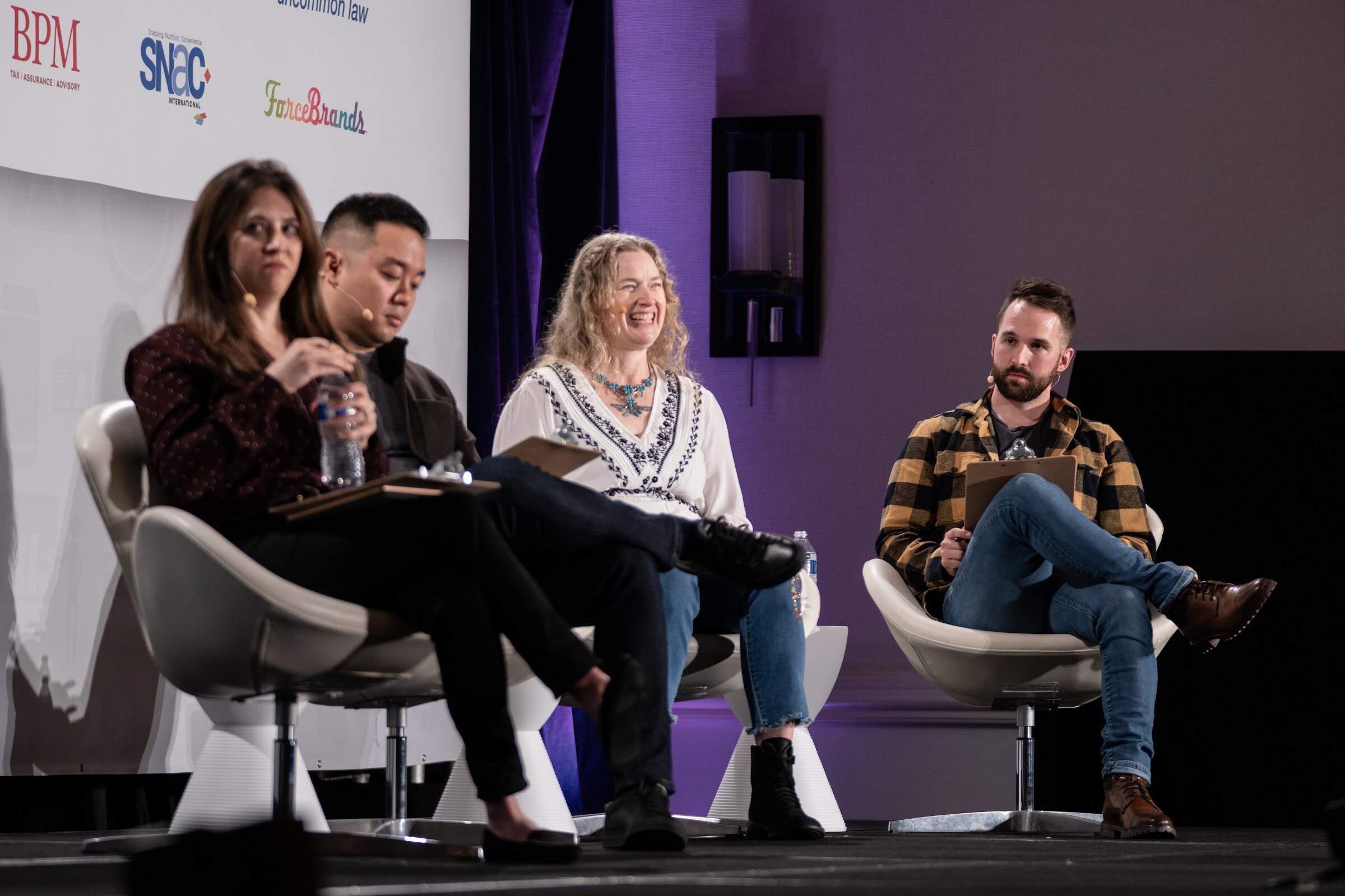 Four people seated on white chairs on a stage, including a woman in a white embroidered blouse and a bearded man in a yellow and black plaid shirt, in front of BPM, SNAC and ForceBrands logos.