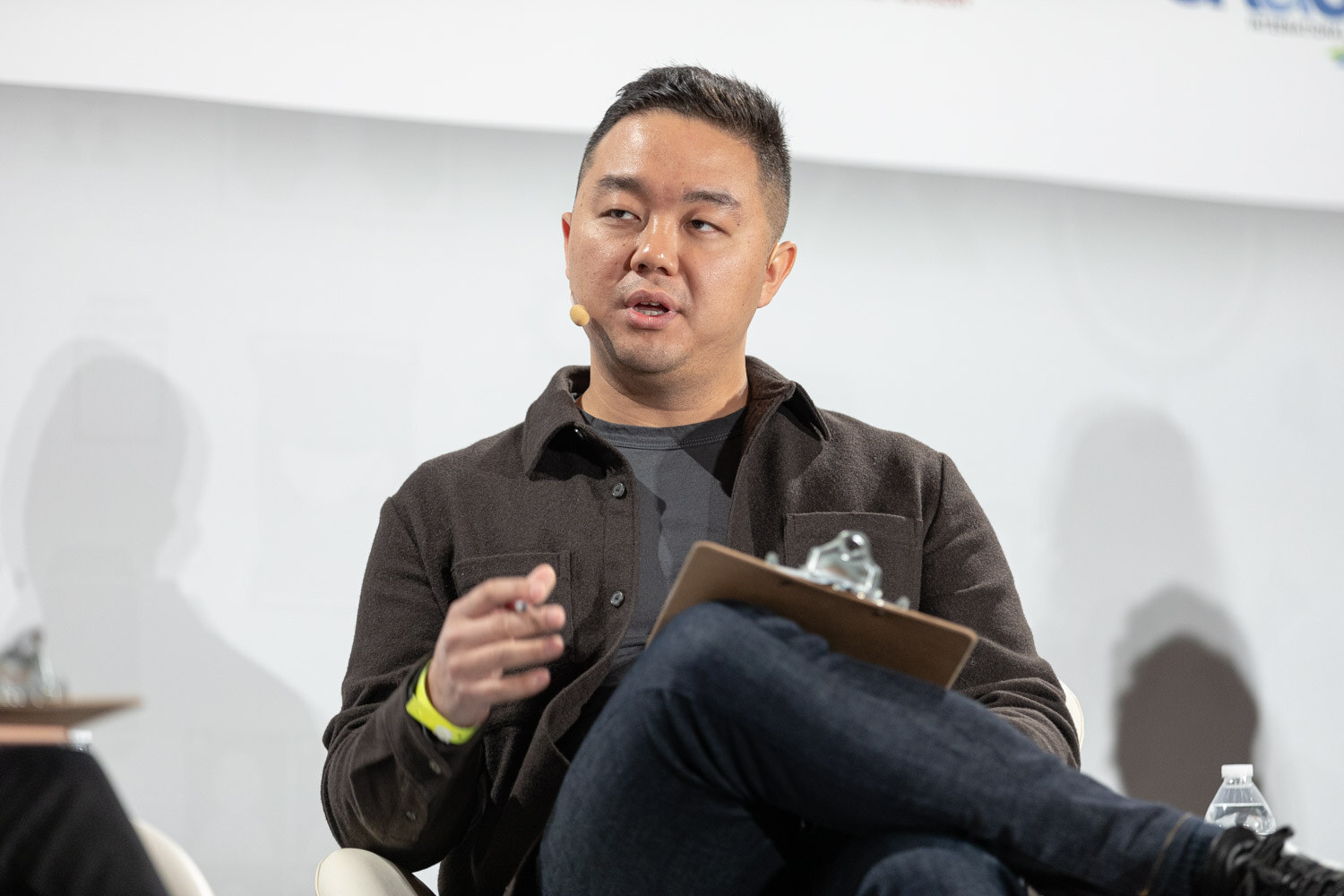 Man in a dark brown overshirt with a headset microphone, a clipboard on his crossed leg and a pen in his hand, in front of a white sponsor backdrop.