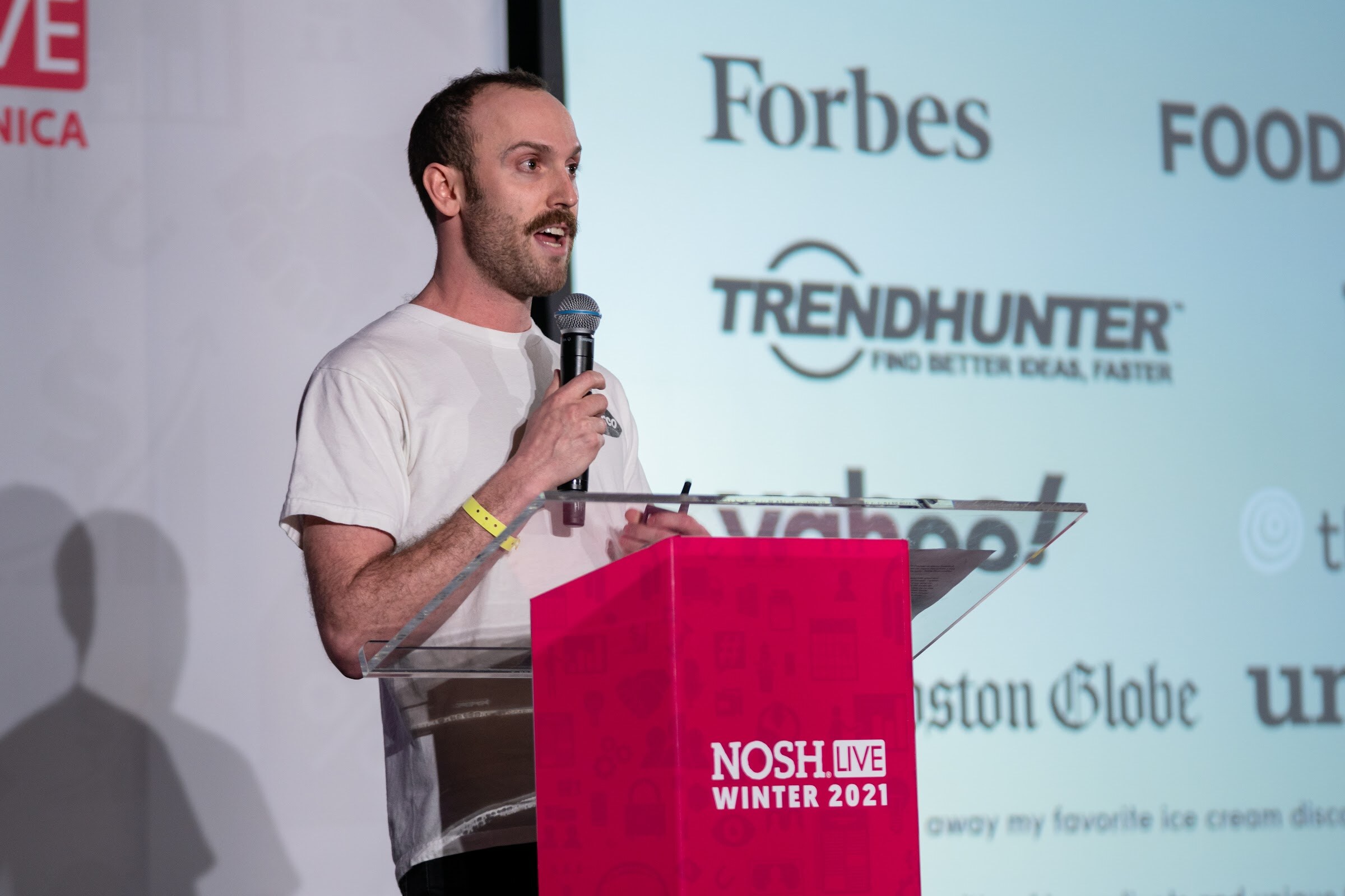 Man in a white t-shirt speaking into a microphone at a pink Nosh Live Winter 2021 podium in front of a slide with Forbes, Trendhunter and Yahoo logos.